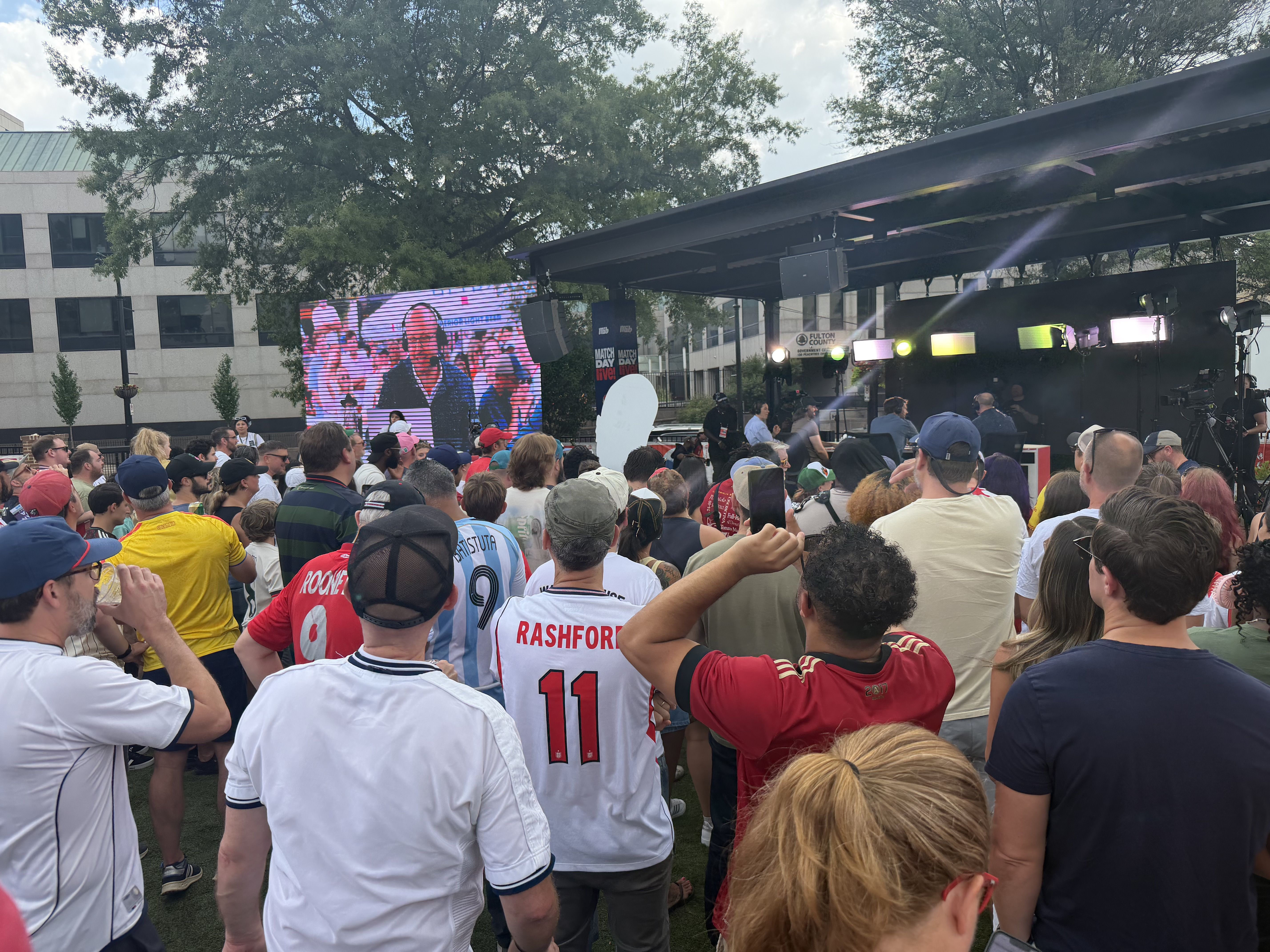 Outdoor event with a right-stage setup, LED screens and cameras. A dense crowd in white Manchester United jerseys (Rashford 11) faces the stage; green trees and a modern building in the background.