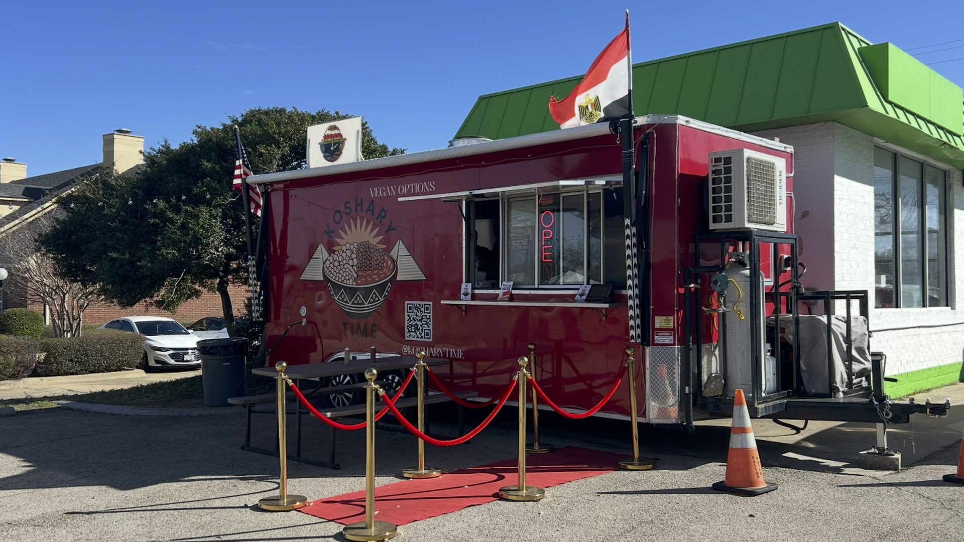 The Koshary Time food truck sits in front of a gas station. The red truck has an Egyptian flag flying over it and a red carpet with red velvet ropes leading to the ordering window. 