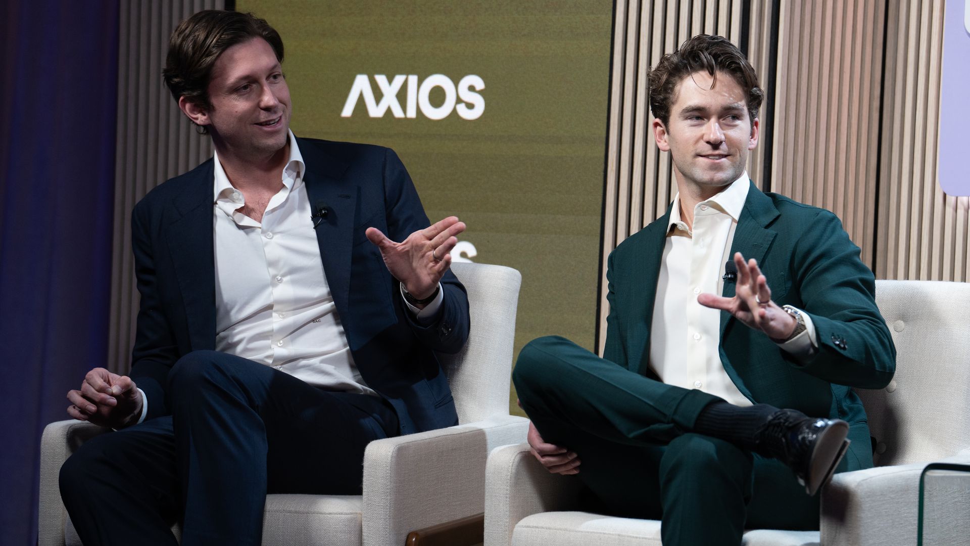 Two men in suits, one navy and one dark green, sit and gesture during a discussion on white armchairs in front of a backdrop with the word "AXIOS".
