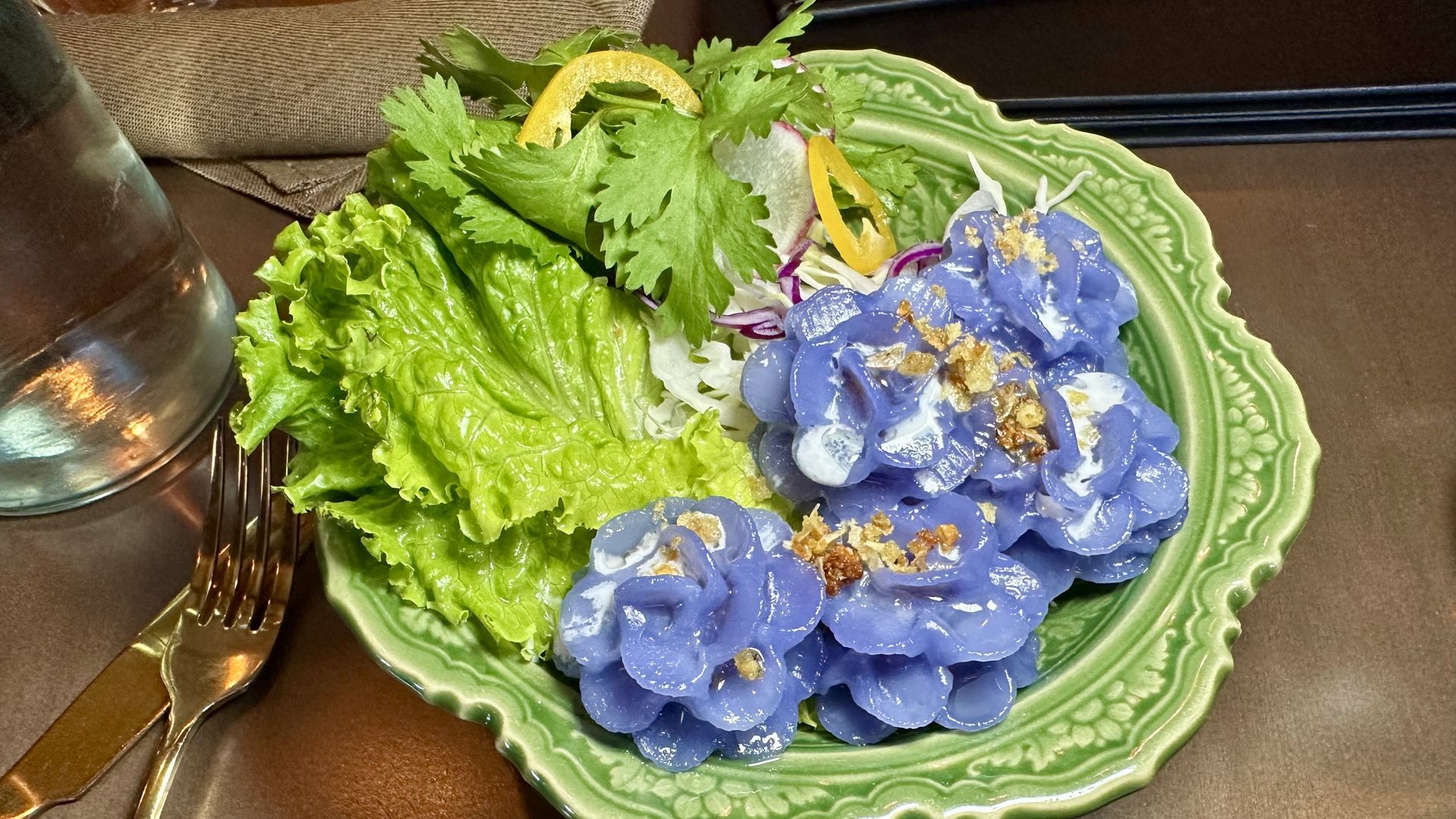 Bright purple dumplings on a green decorative plate, topped with golden flecks and surrounded by lettuce, cilantro, and lemon zest; a fork and knife rest on a brown table beside a glass.