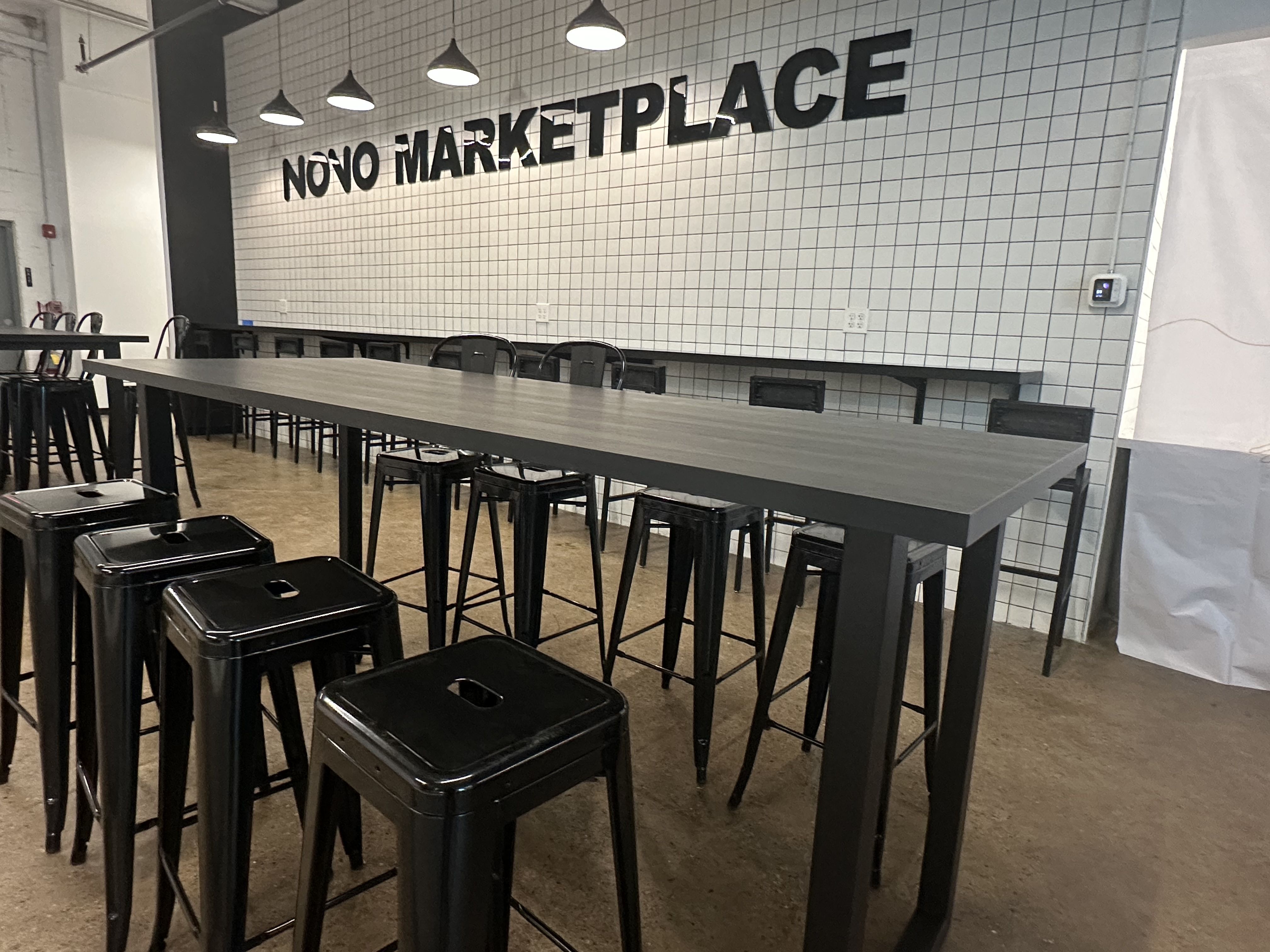 Interior of a modern market hall with a white grid-tile wall reading the bold text "NÒVO MARKETPLACE". Long gray tables with black stools under industrial pendant lights.