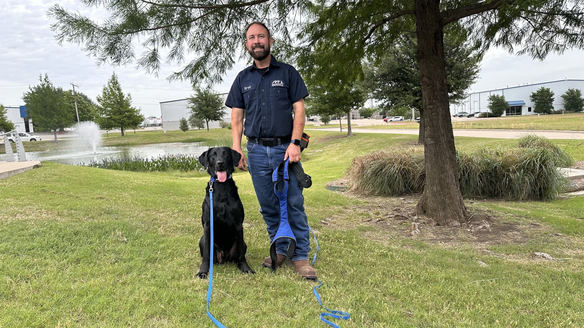 A photo of River, a black labrador retriever mix, and his human coworker Brad Blan standing outside under a tree in front of a water feature in Mansfield, Texas.