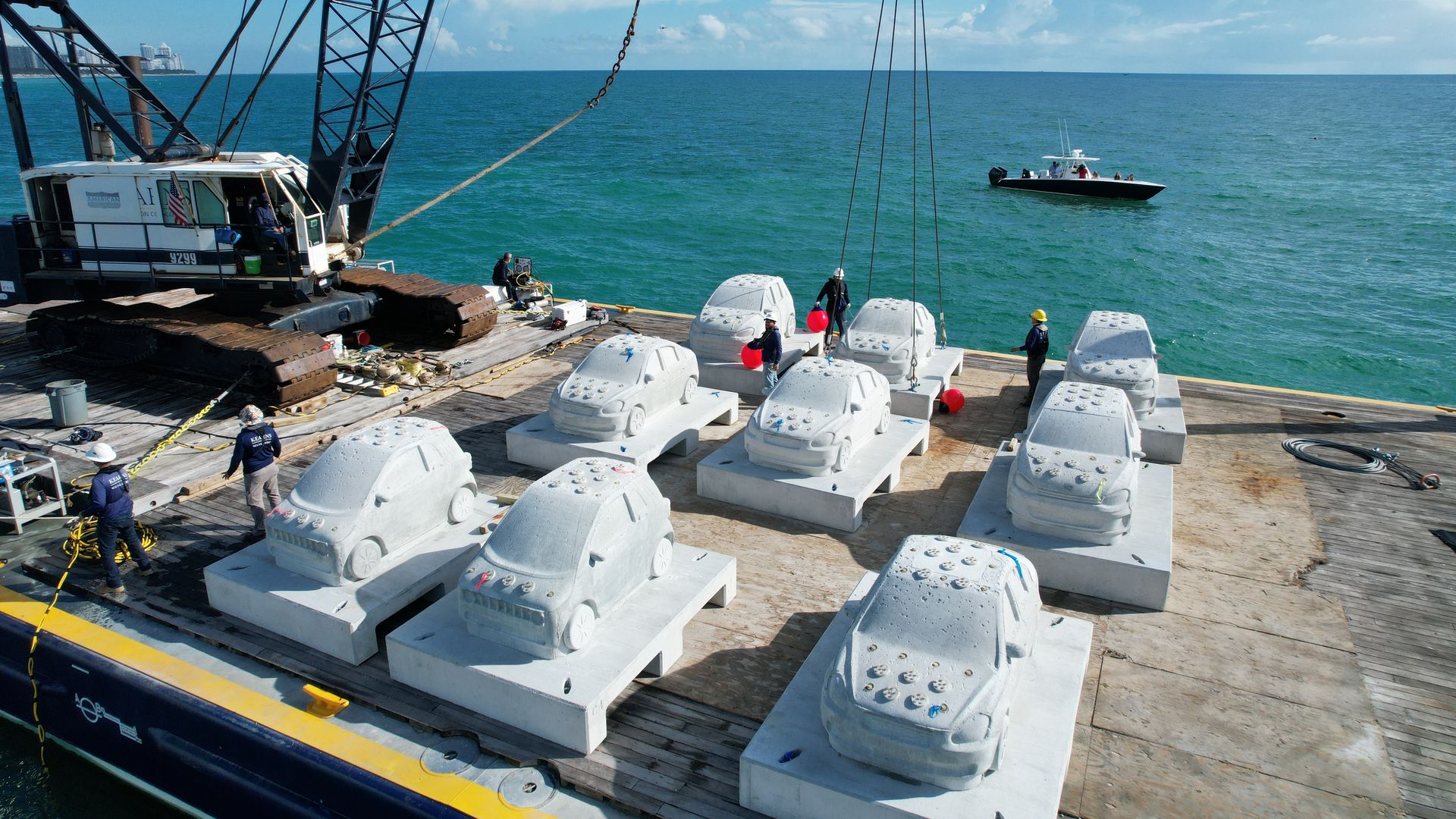 Large crane on a barge lifts concrete car-shaped structures over the ocean with workers nearby and a small boat in the distance under a blue sky with clouds.
