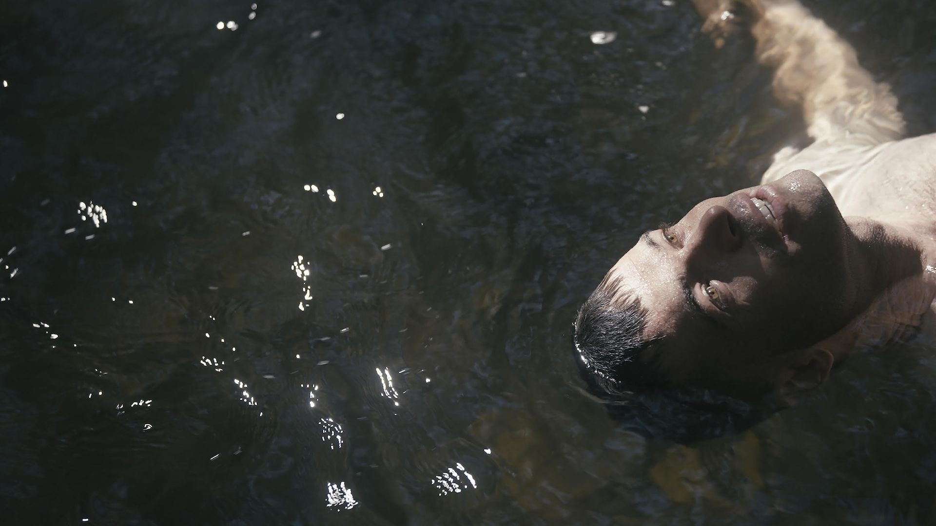 A man with wet dark hair floats on his back in dark water, his eyes open and light reflecting on the rippling surface around him.