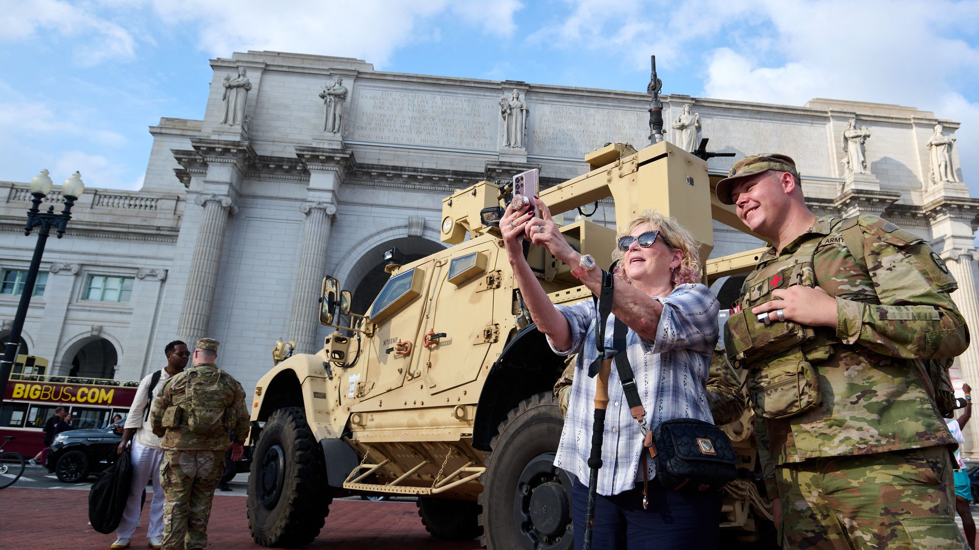 A woman wearing sunglasses takes a selfie with a smiling soldier in camouflage uniform standing beside a large tan military vehicle in front of a grand white stone building under a blue sky.