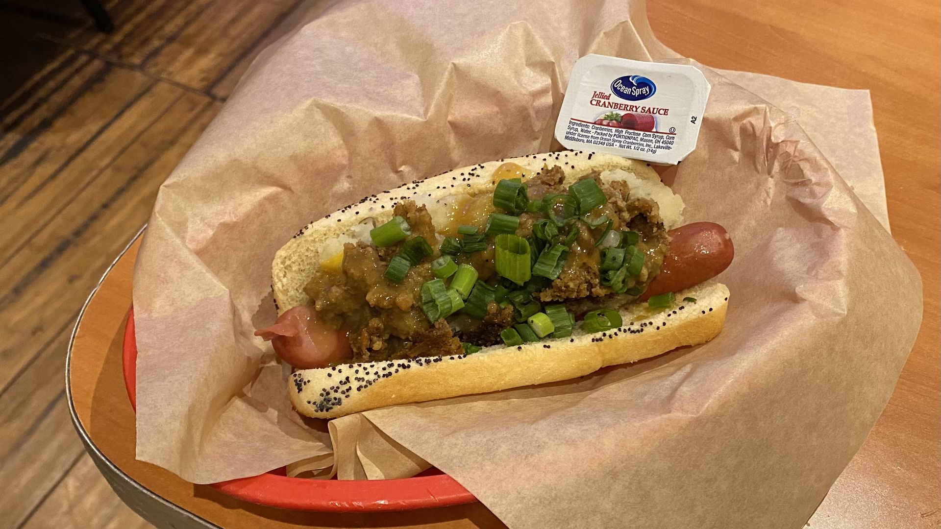 A hot dog in a basket topped with stuffing, gravy, mashed potatoes and scallions, with a side of prepackaged cranberry sauce