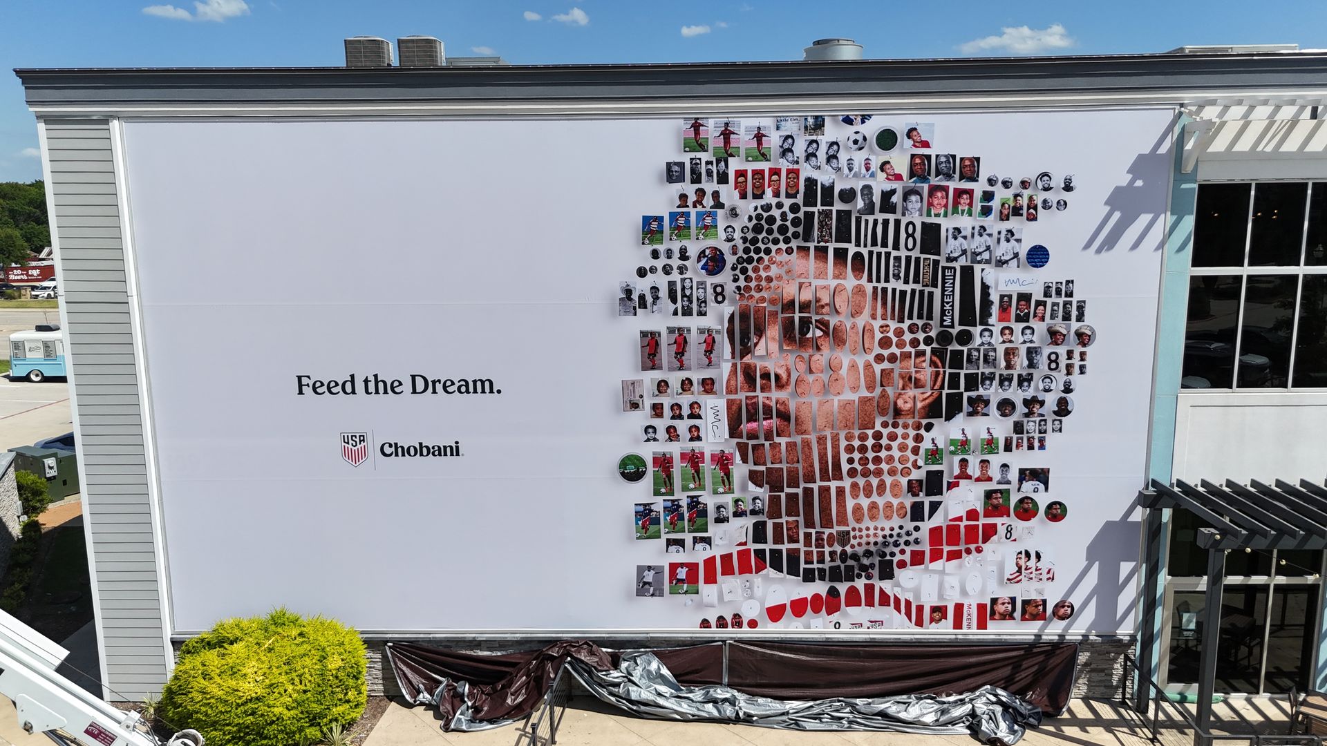 Exterior wall mural on a building featuring a dense mosaic of small portraits and circular images forming a large pattern; left side reads "Feed the Dream" with "USA Chobani" logo, blue sky above.