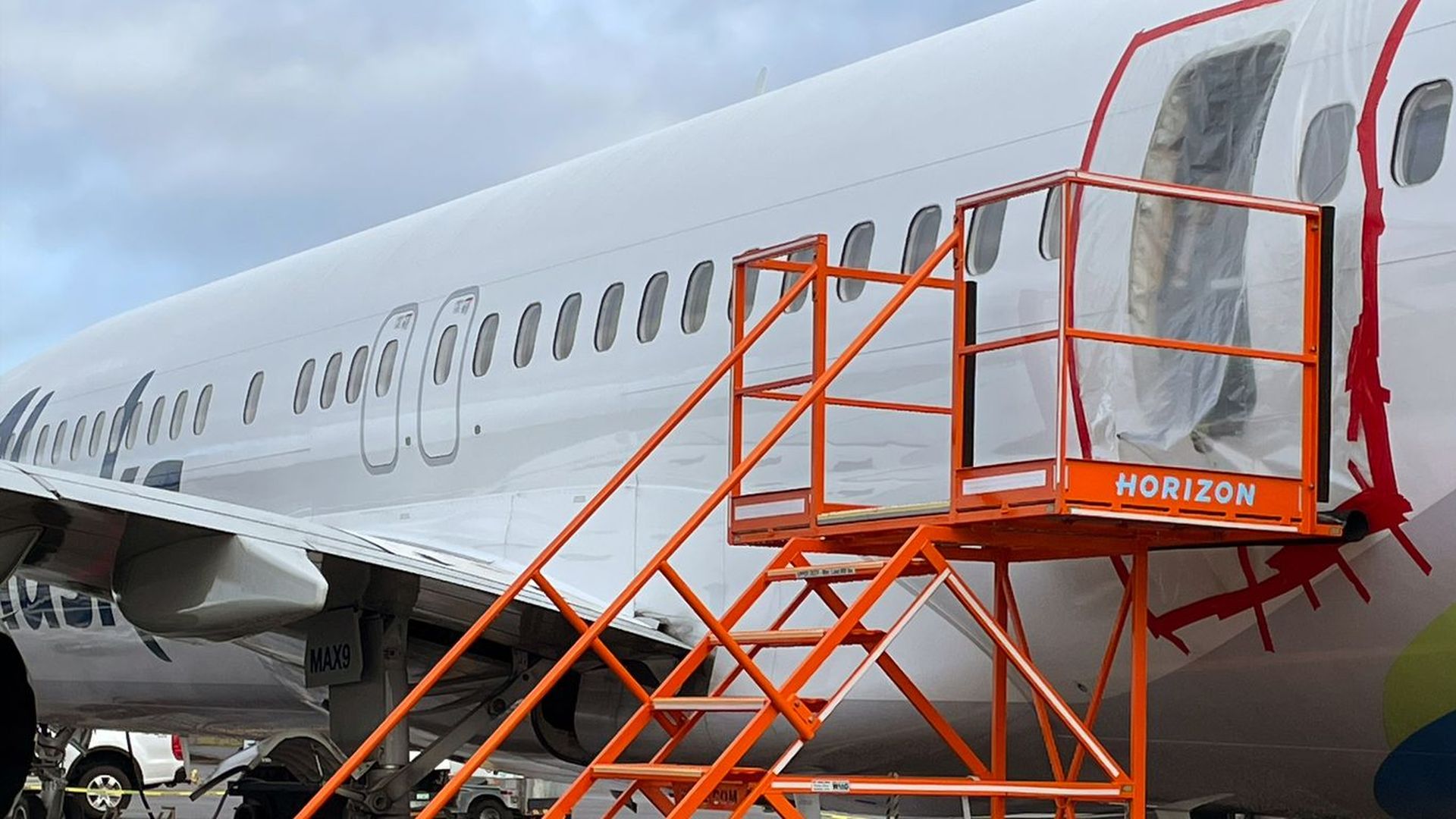 An airplane with an orange stair cart propped up against an open doorway with a plastic sheet draped over it.