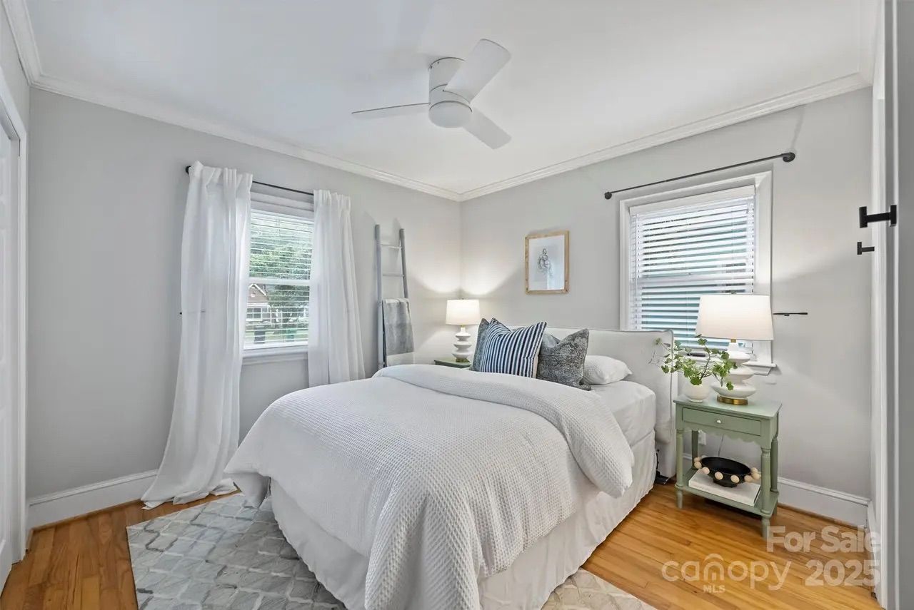 Bright bedroom with white walls, hardwood floor, ceiling fan, white bed with blue and patterned pillows, green nightstand with lamp and plant, two windows with white curtains.