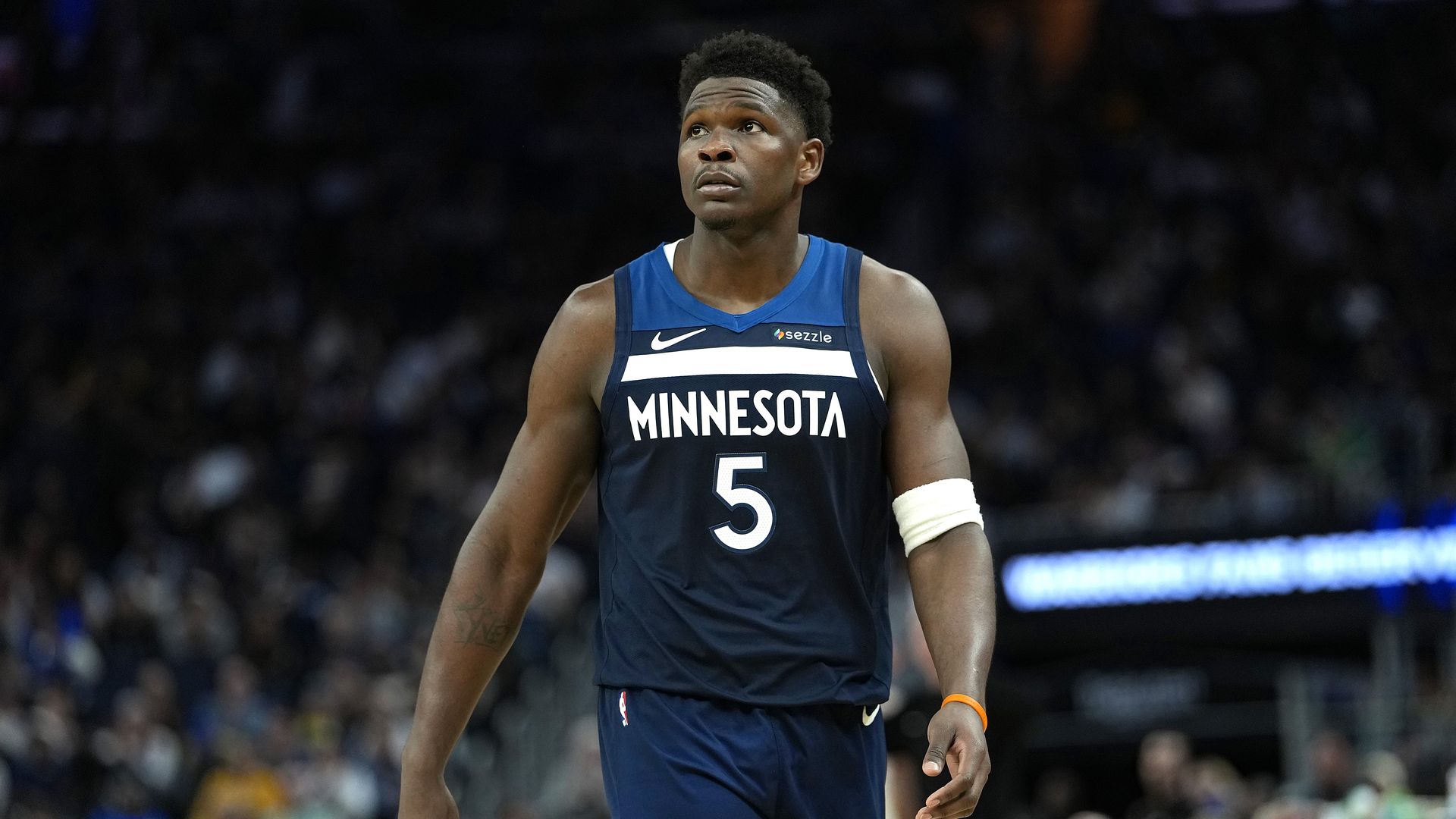 Anthony Edwards a dark blue Minnesota jersey with the number 5 and an orange wristband, on a court with a blurred crowd.