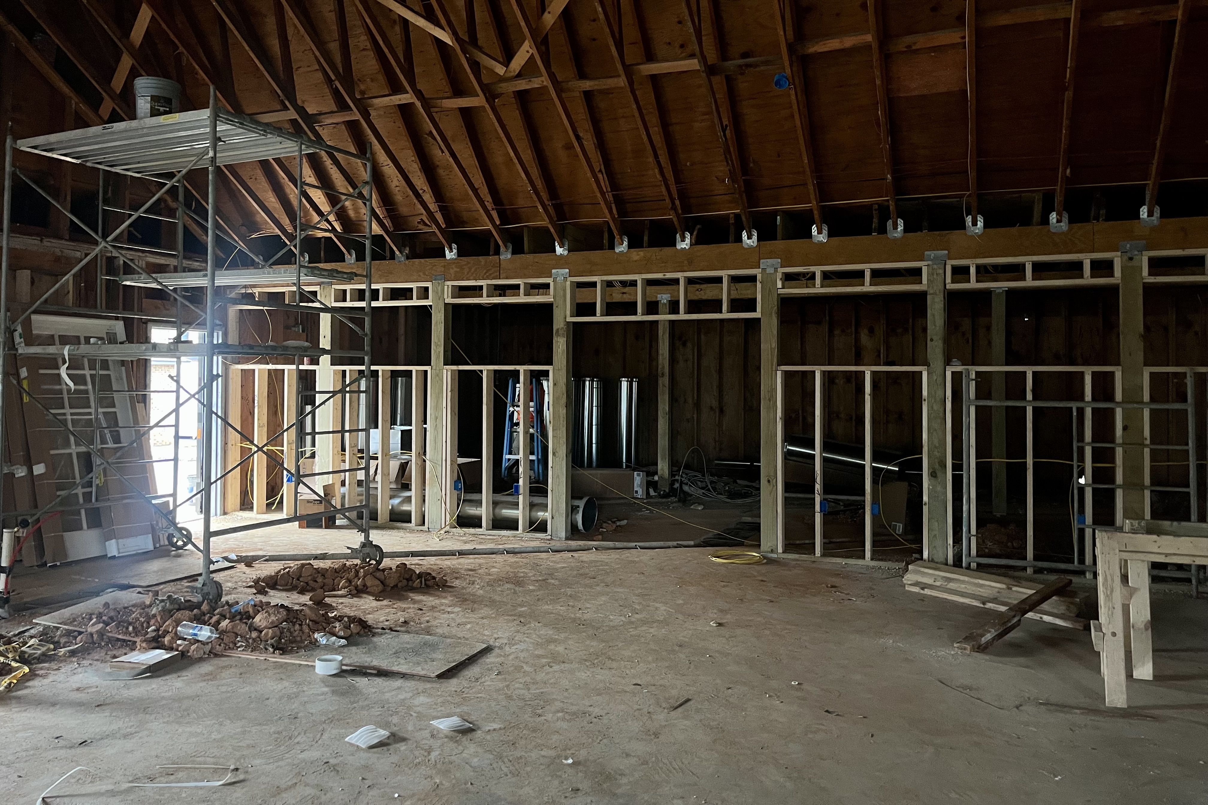 Interior of a building under construction with exposed wooden framing, metal scaffolding, dirt floor scattered with tools and debris, and a wooden vaulted ceiling.