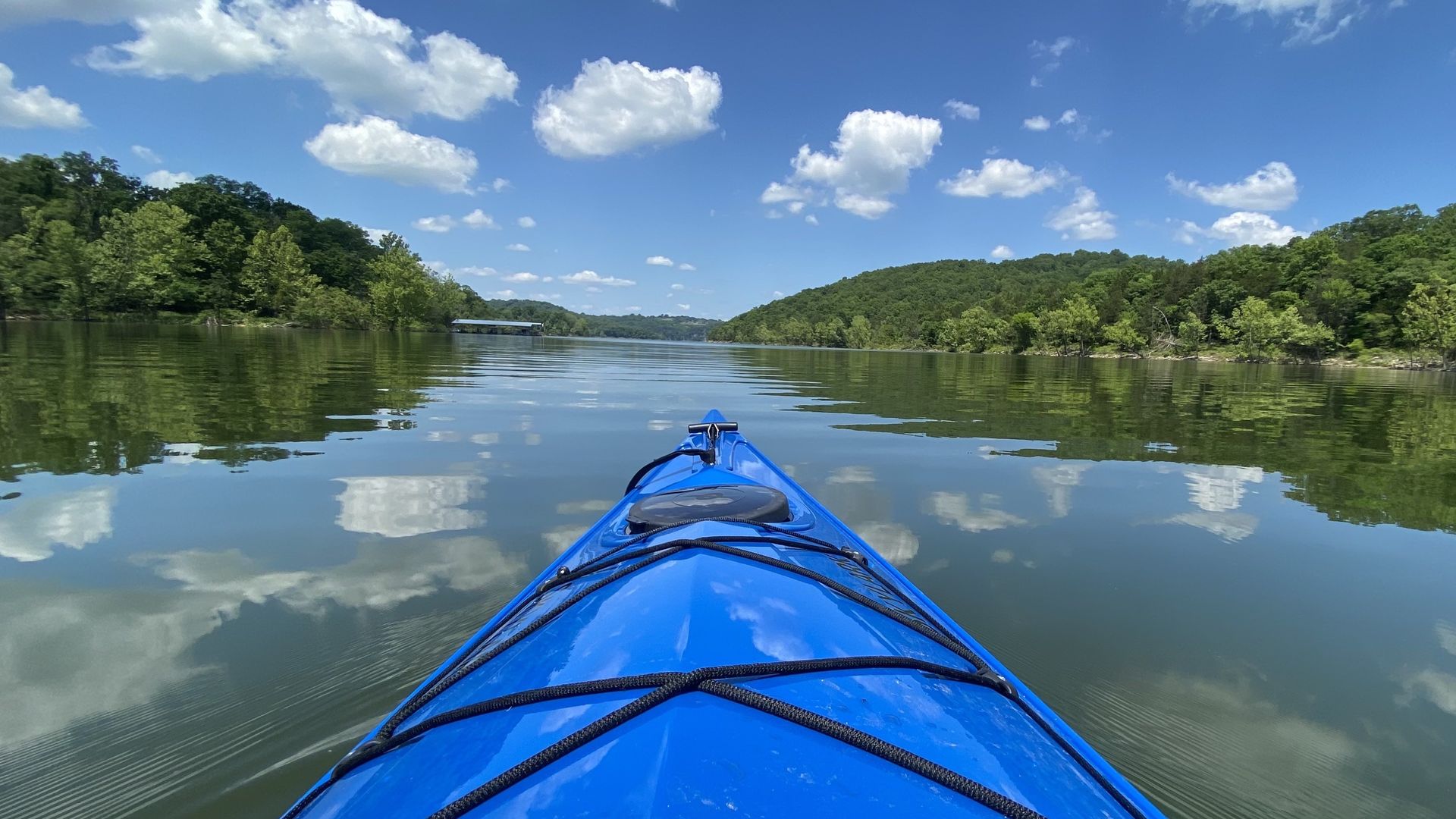 A photo of the front of a blue kayak on water reflecting the clouds in the sky, framed with greenery