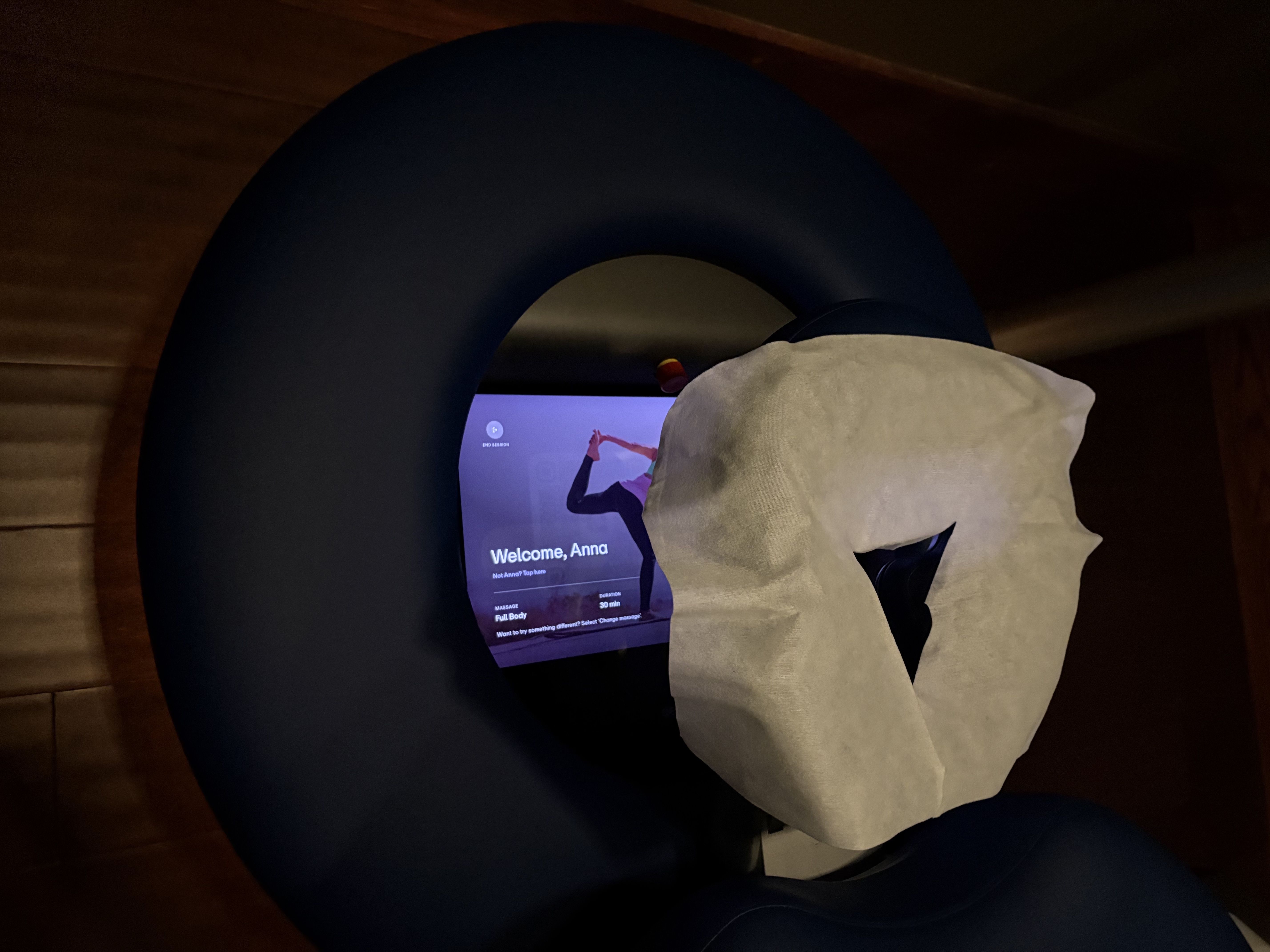 Dark blue massage chair with a face cradle covered by white tissue paper, a screen displays a welcome message for Anna and options for a 30-minute full body massage.