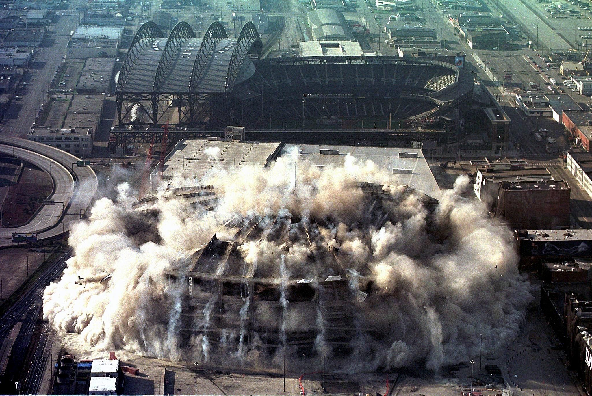 The Kingdome begins to collapse, with dust spreading out as the concrete falls to the ground.