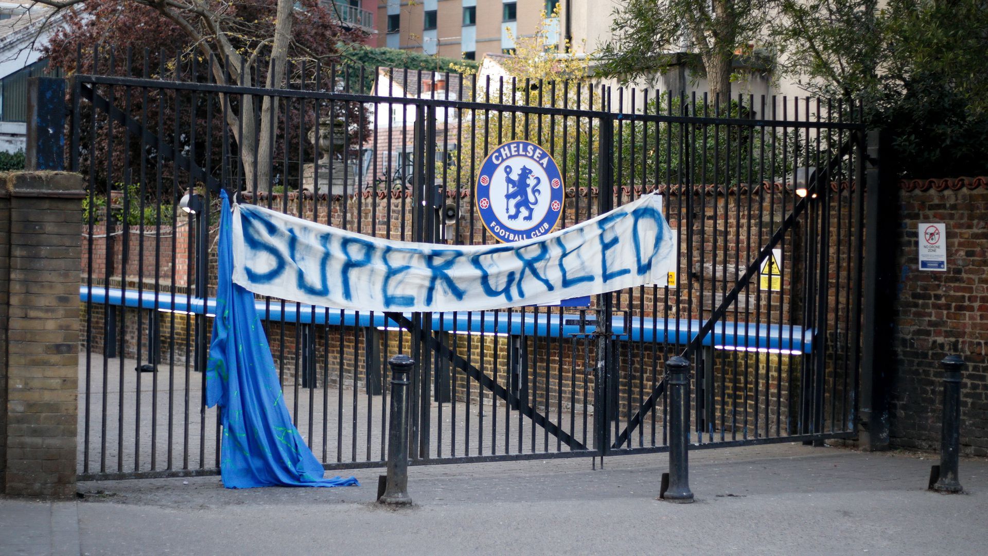 A sign reading "Super greed" hangs on a fence with the chelsea football club logo.
