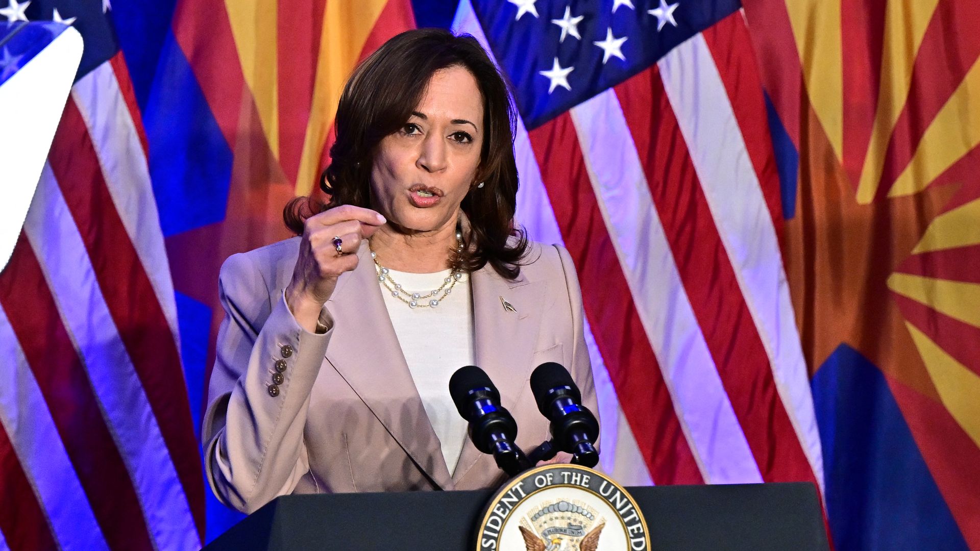 A woman speaks into microphones while standing in front of American and Arizona flags.