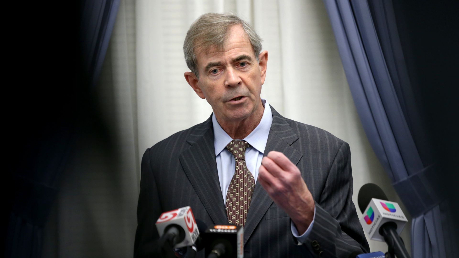 Massachusetts Secretary of State Bill Galvin, a middle-aged man in a dark pinstripe suit and patterned brown tie, speaking at a press conference with multiple microphones, framed by blue curtains and a white background.