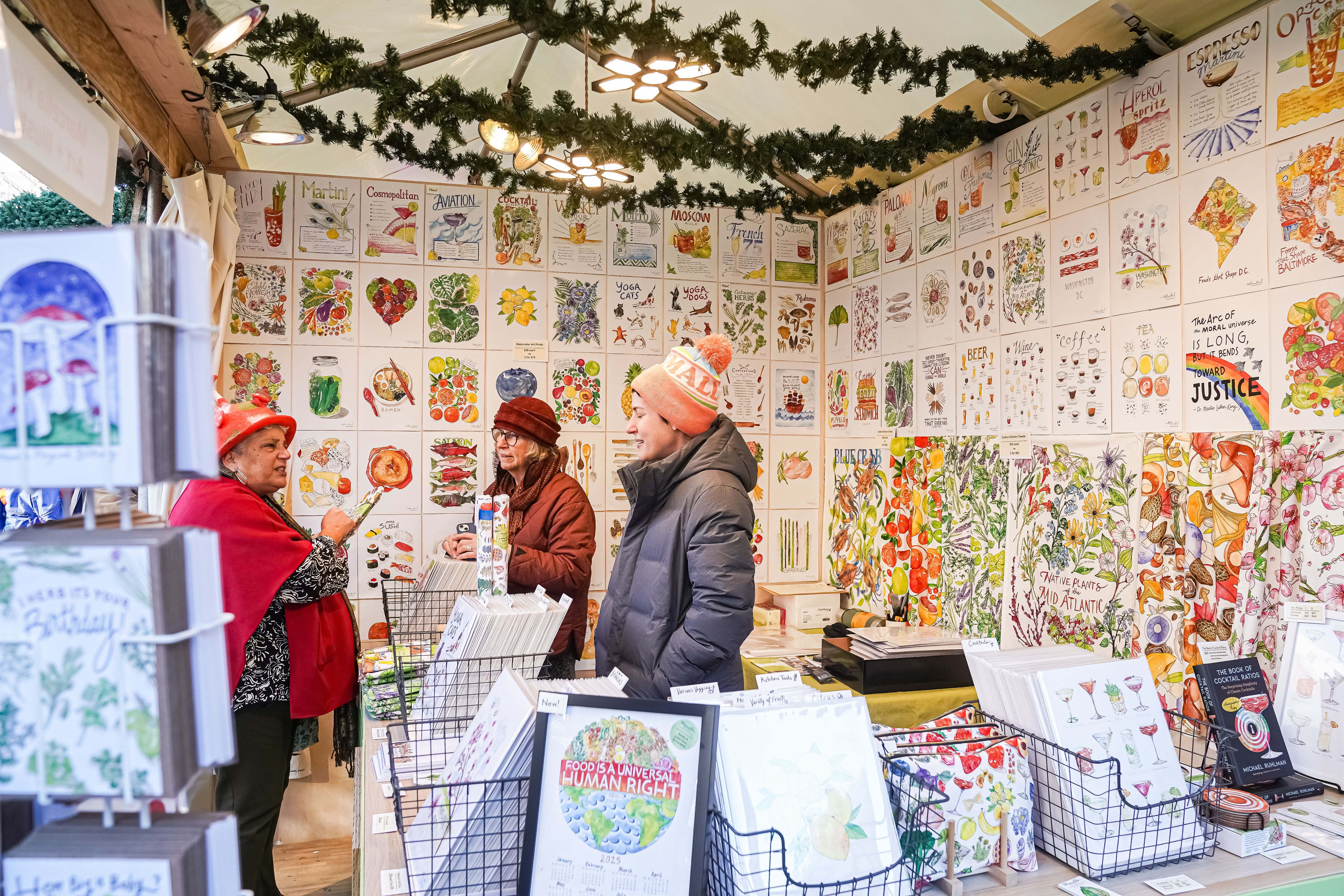 Three people in warm clothing inside a booth decorated with colorful illustrated posters of food, drinks, plants, and quotes, with garlands hanging from the ceiling.