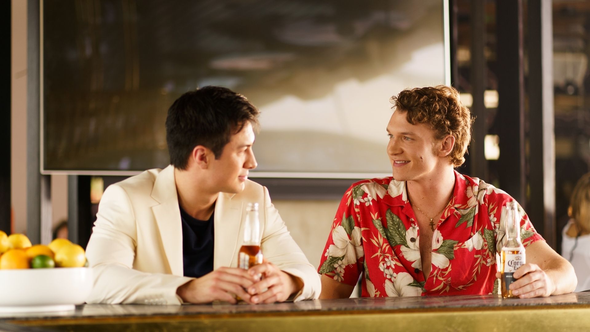 Two young men smiling at each other while sitting at a bar, one wearing a white blazer, the other a red floral shirt, each holding a bottle of Corona beer.