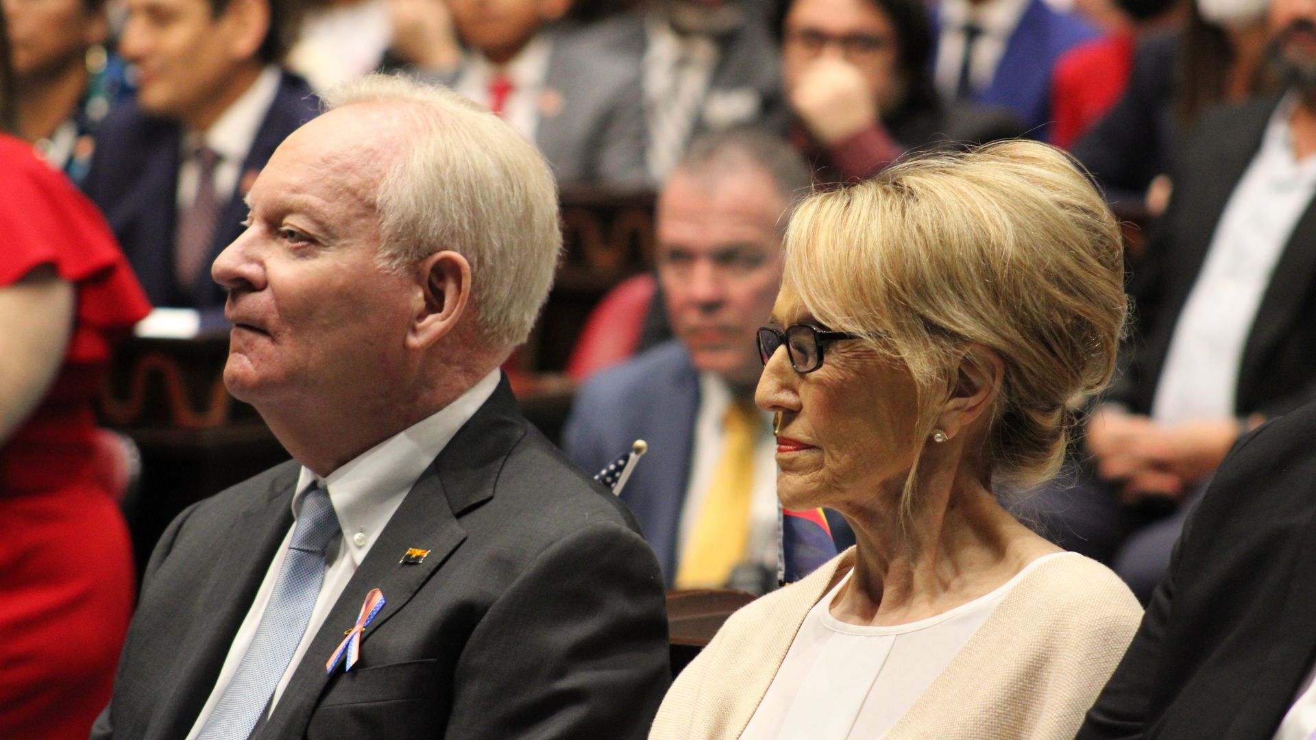A man and a woman sit next to each other with a crowd behind them.