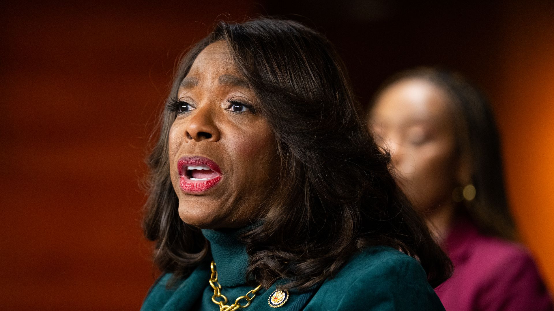 Close-up of a Black woman speaking, wearing teal attire with a gold chain and lapel pin, pink lipstick and dark hair; warm orange background with a blurred person behind.