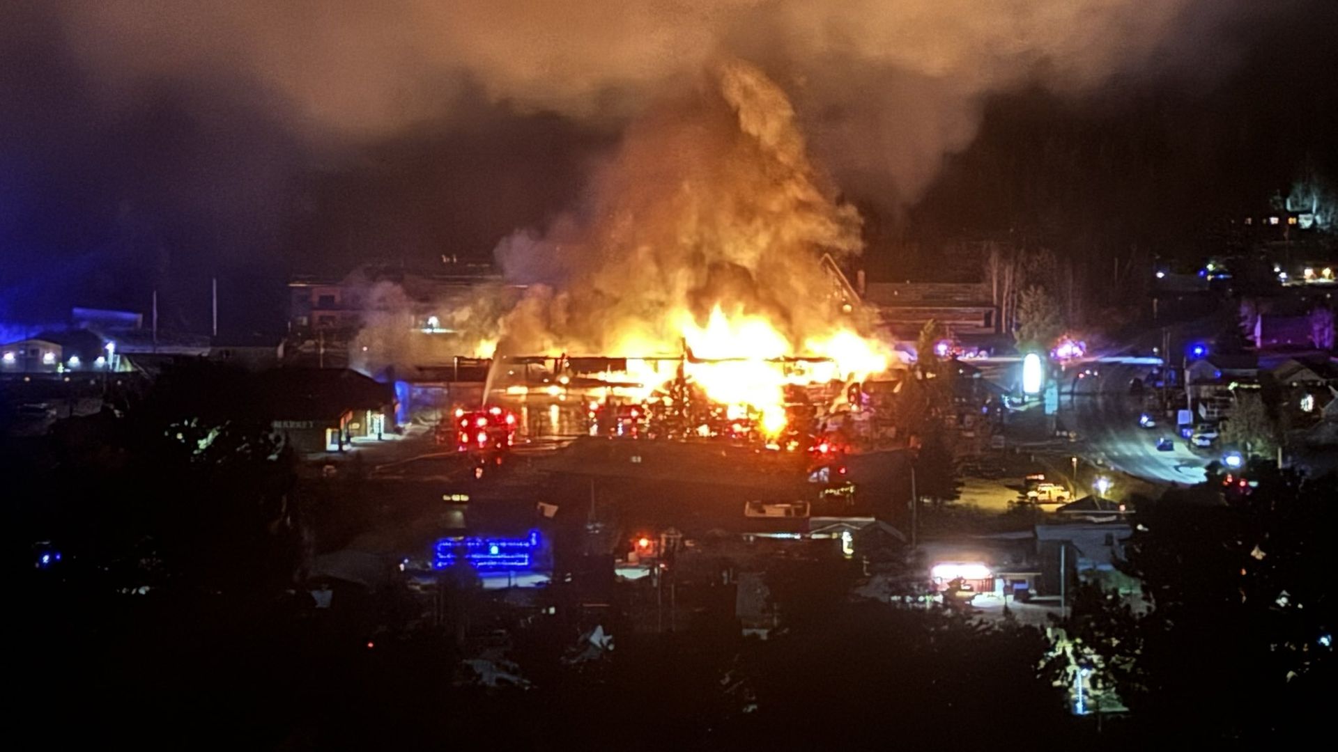 Large building engulfed in bright flames and smoke at night with emergency vehicles and flashing lights surrounding the scene in a town area.