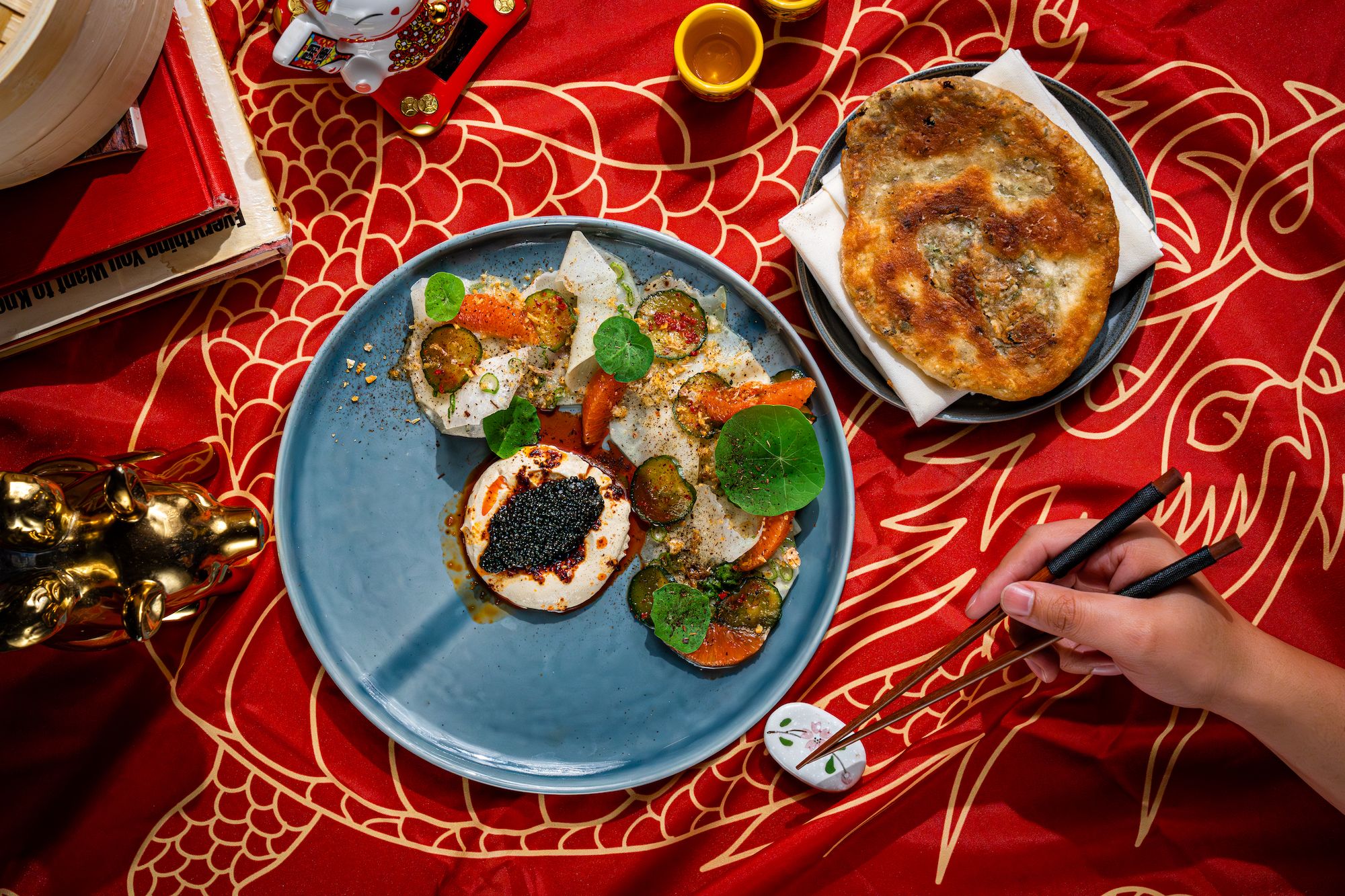 A red dragon tablecloth set with a blue ceramic plate with caviar and whipped tofu, plus a separate plate with a scallion pancake
