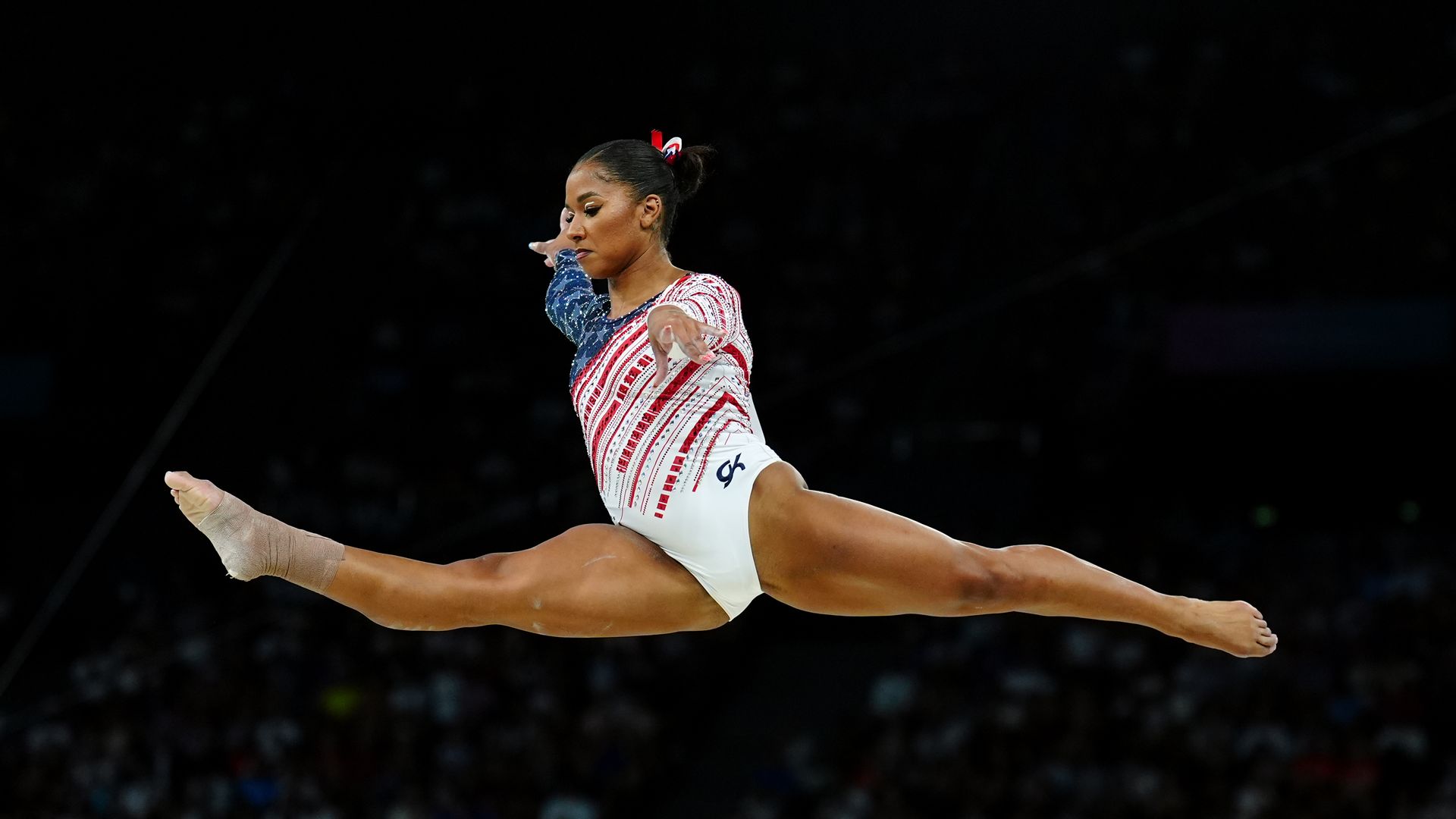 Jordan Chiles leaps in midair with one leg straight ahead of her and one straight behind, arms out, wearing the Team USA gymnastics uniform.