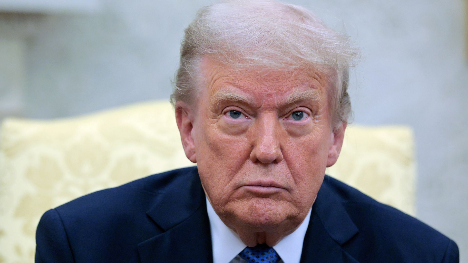 President Donald Trump faces the camera while sitting in a high-back yellow chair in the Oval Office. 