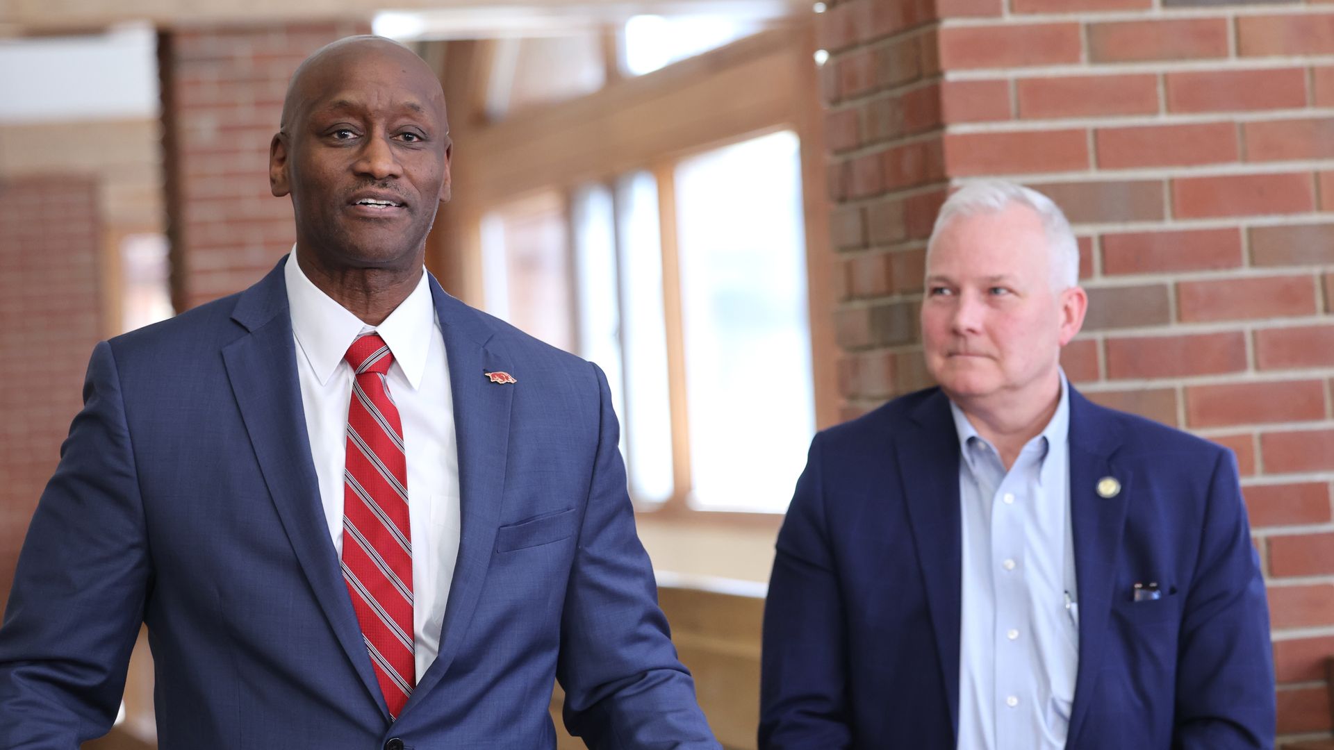 Two men in blue suits standing indoors by brick wall; man on left wears red and white striped tie, man on right has light blue shirt and no tie.