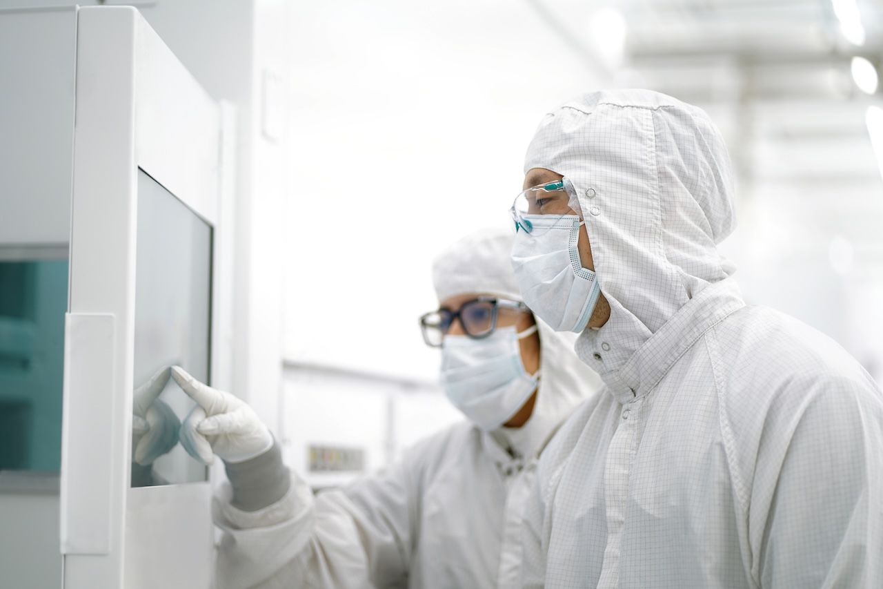 Two scientists in white cleanroom suits, masks, and goggles operate a large white lab machine; one touches a touchscreen while the other watches.