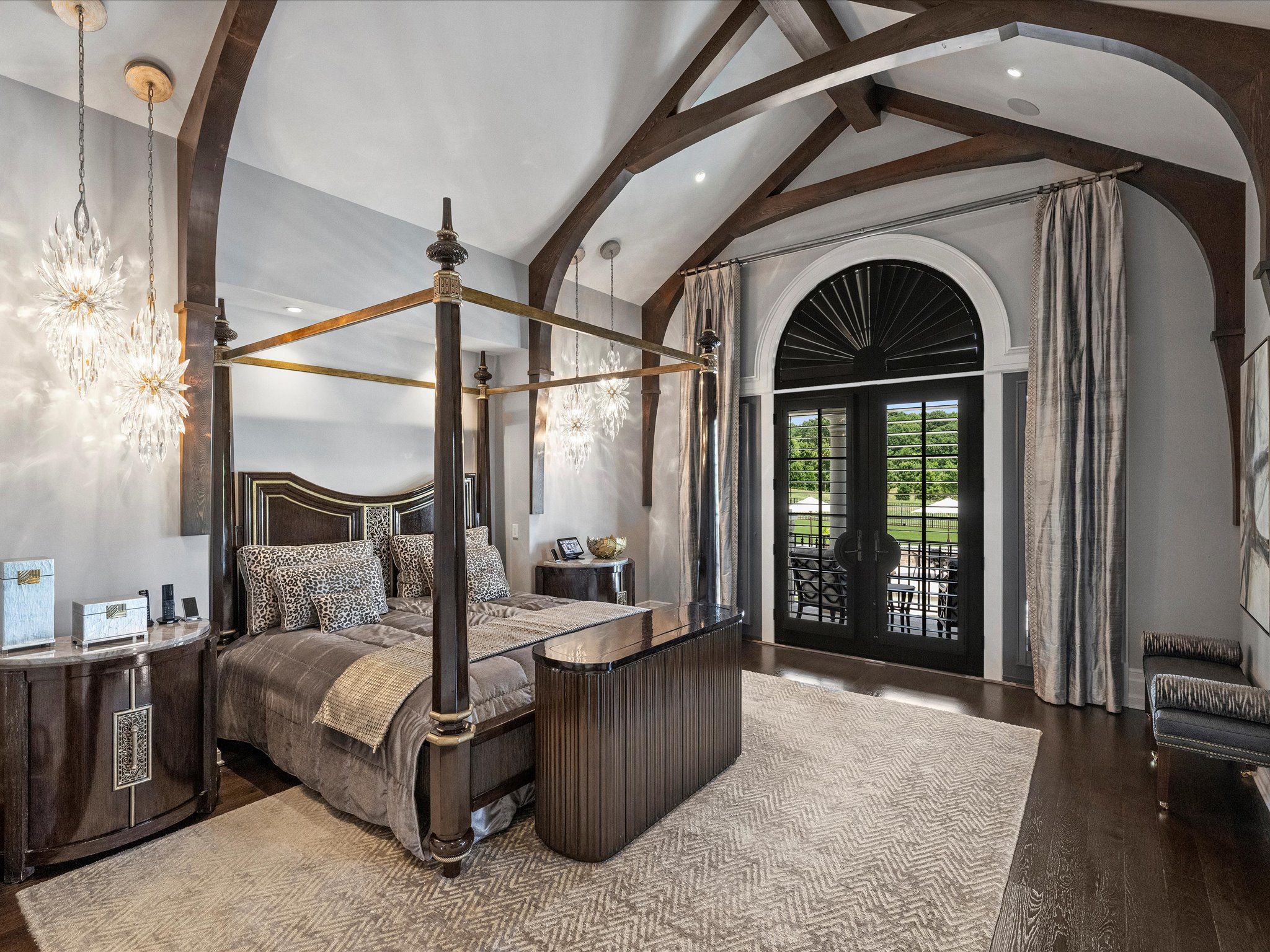 Elegant bedroom with dark wooden four-poster bed, leopard print pillows, beige rug, arched black double doors with shutters, gray curtains, and crystal pendant lights. Dark wooden beams on ceiling.