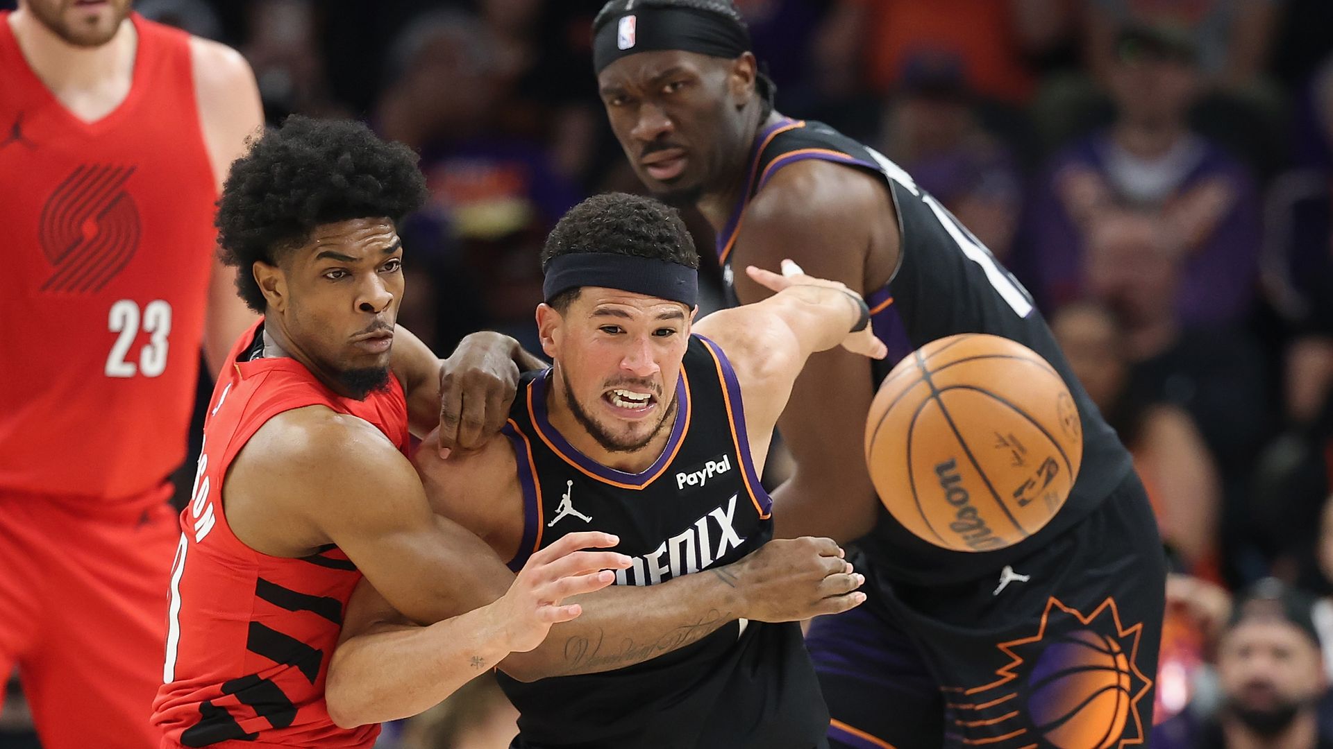 A basketball player in a red uniform holds back a player in a black uniform who's trying to reach a loose ball, with another player from each team in the background. 