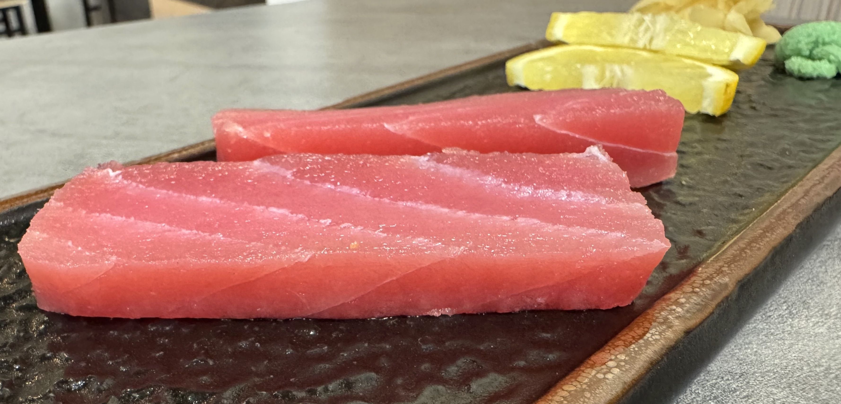 Two thick slices of raw tuna sashimi are served on a black plate with lemon wedges, pickled ginger, and a small mound of wasabi.