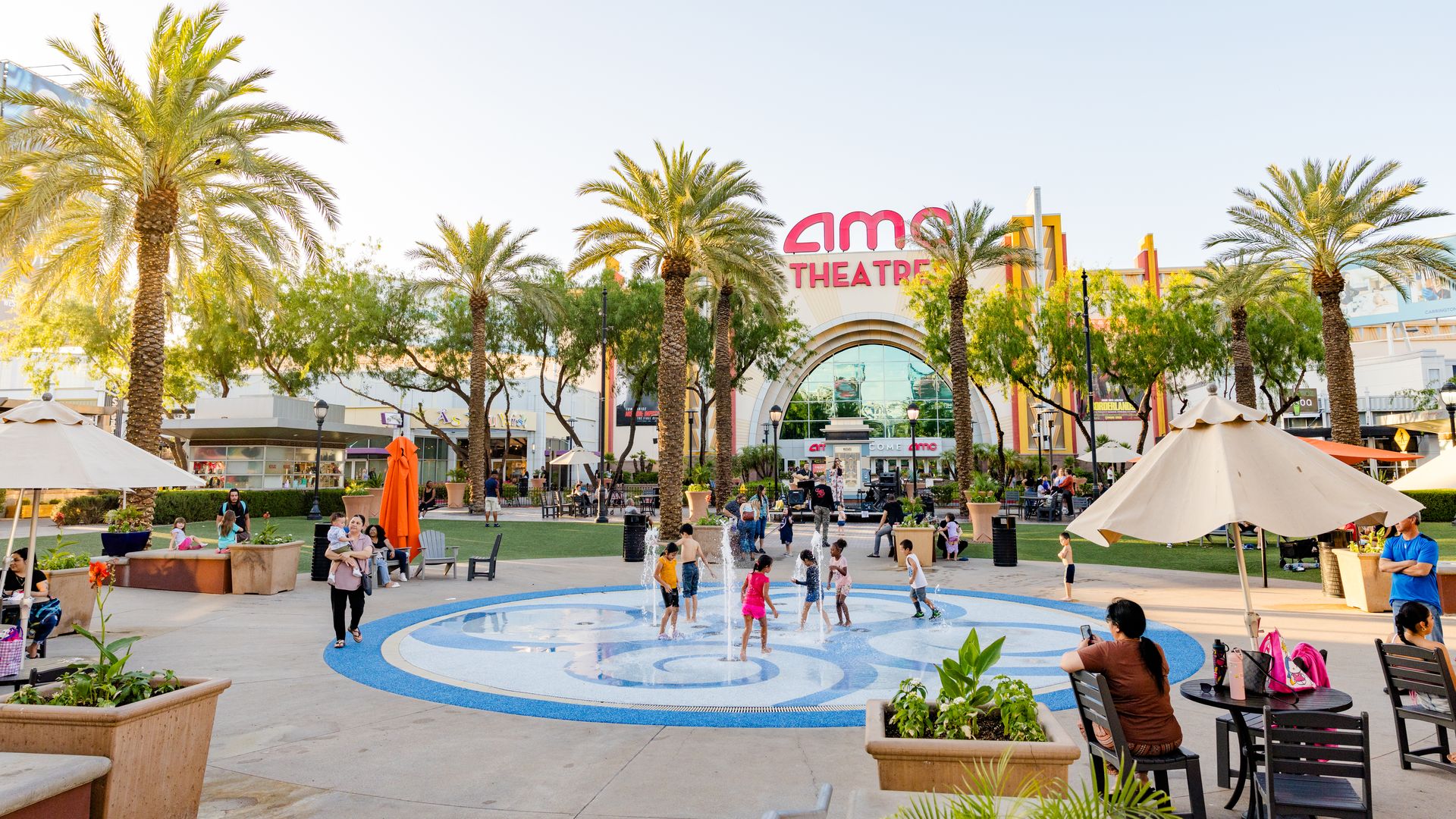 Sunny outdoor plaza lined with palm trees, a circular water fountain, and seating. People stroll and play near the bright AMC Theatre entrance under a clear blue sky.