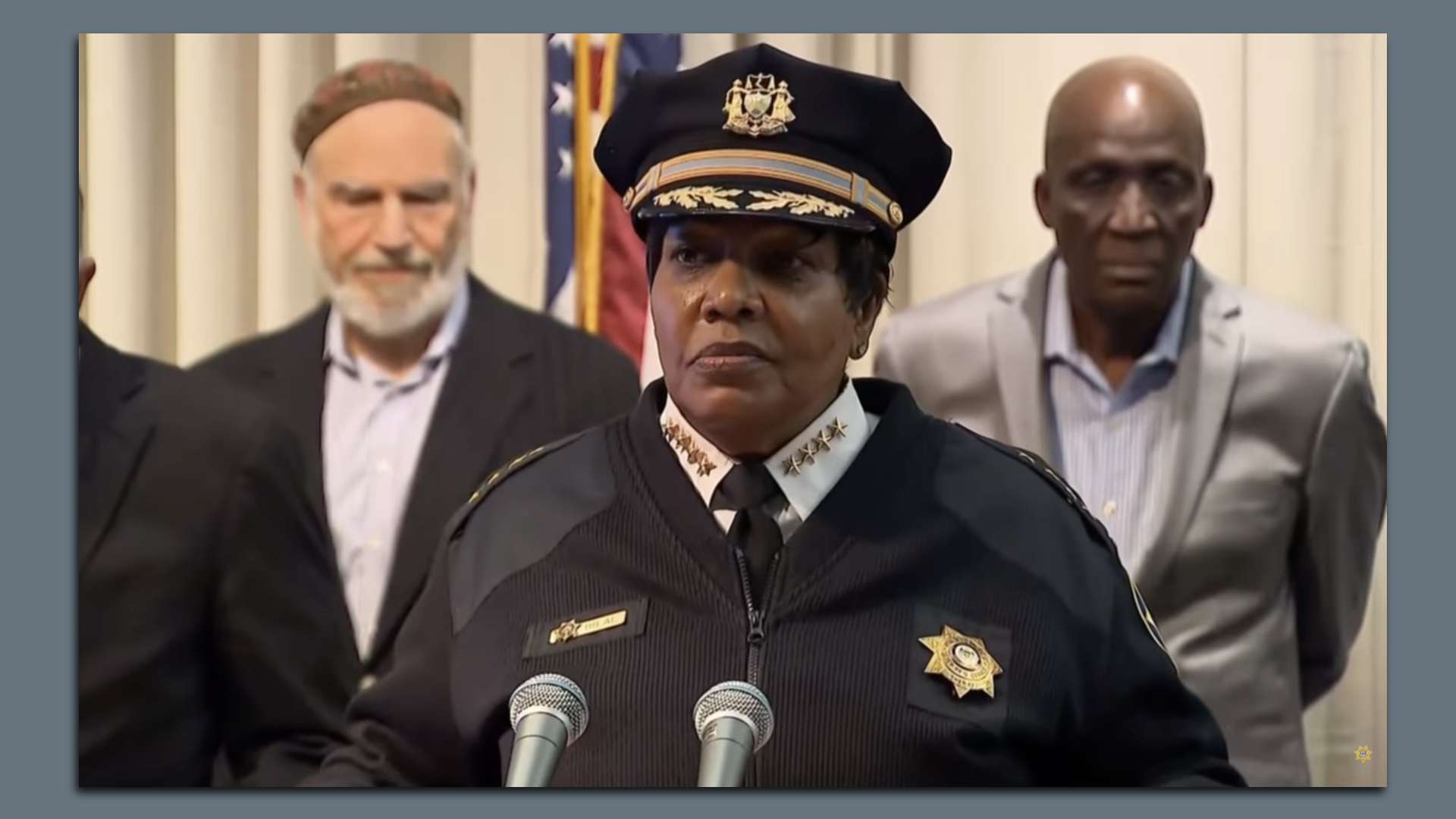 Black female police chief speaking at podium with two men standing behind, American flag in background, indoor setting, serious expressions.