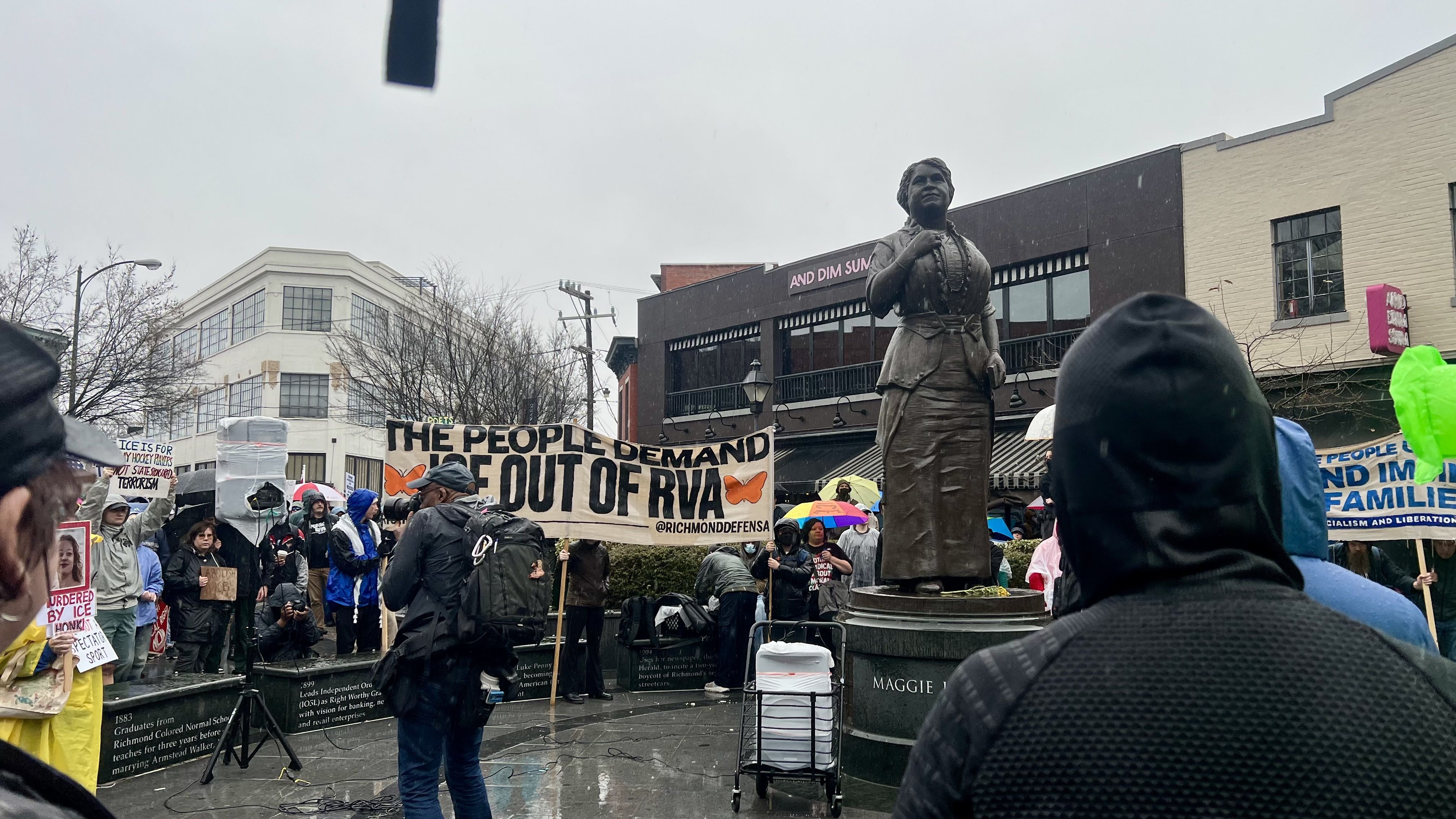 Crowd gathered at rainy protest near bronze statue of woman on pedestal inscribed "Maggie L." People hold banners, including one reading "THE PEOPLE DEMAND ICE OUT OF RVA" with orange butterflies.
