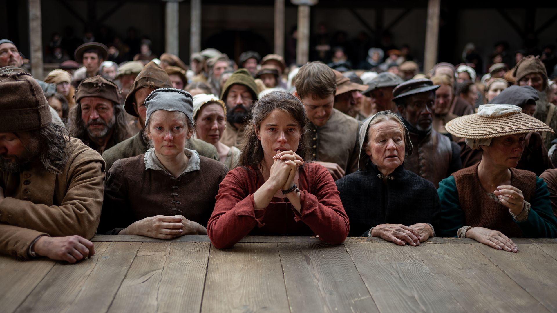 A crowd of people in old, worn clothing stand behind a wooden table, looking somber and anxious, with a young woman in a red dress sitting at the table, clasping her hands in front of her face.