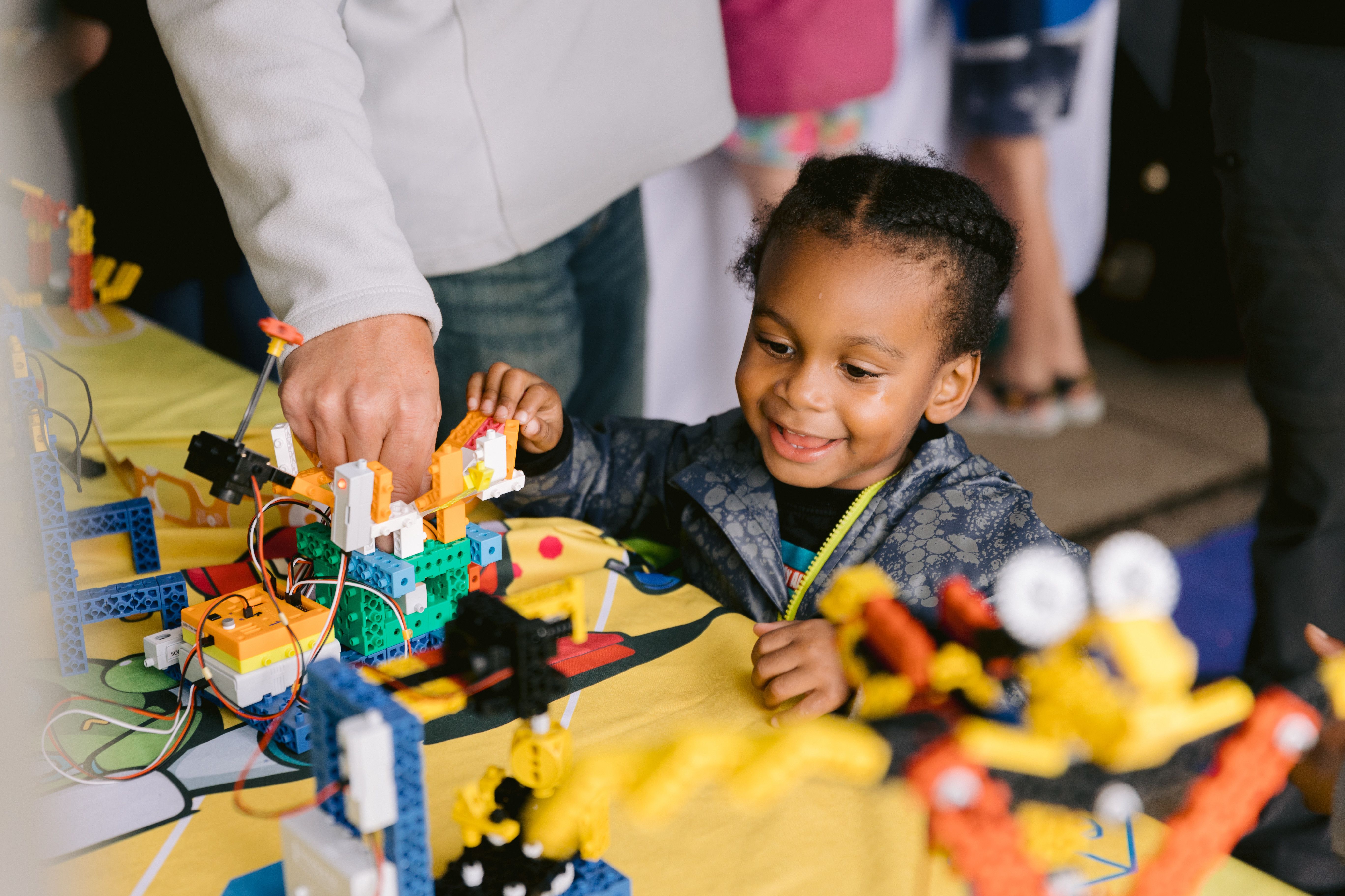 A smiling child builds with colorful LEGO-like bricks on a bright yellow table, guided by an adult's hands; tangled wires and more blocks create a busy, playful construction scene.