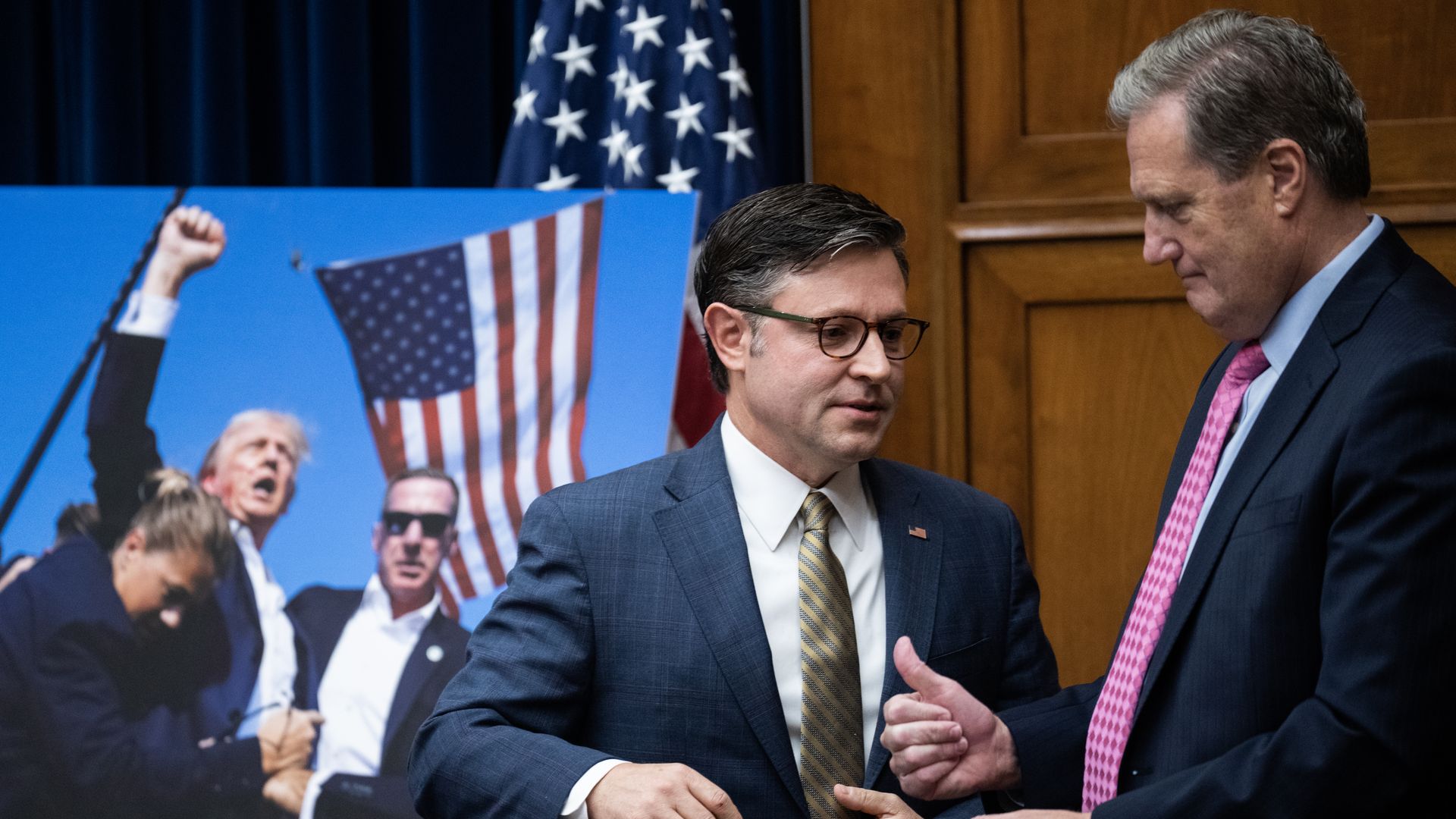 Reps. Mike Johnson and Mike Turner in front of a photo of the first Trump assassination, an American flag and a wooden wall.