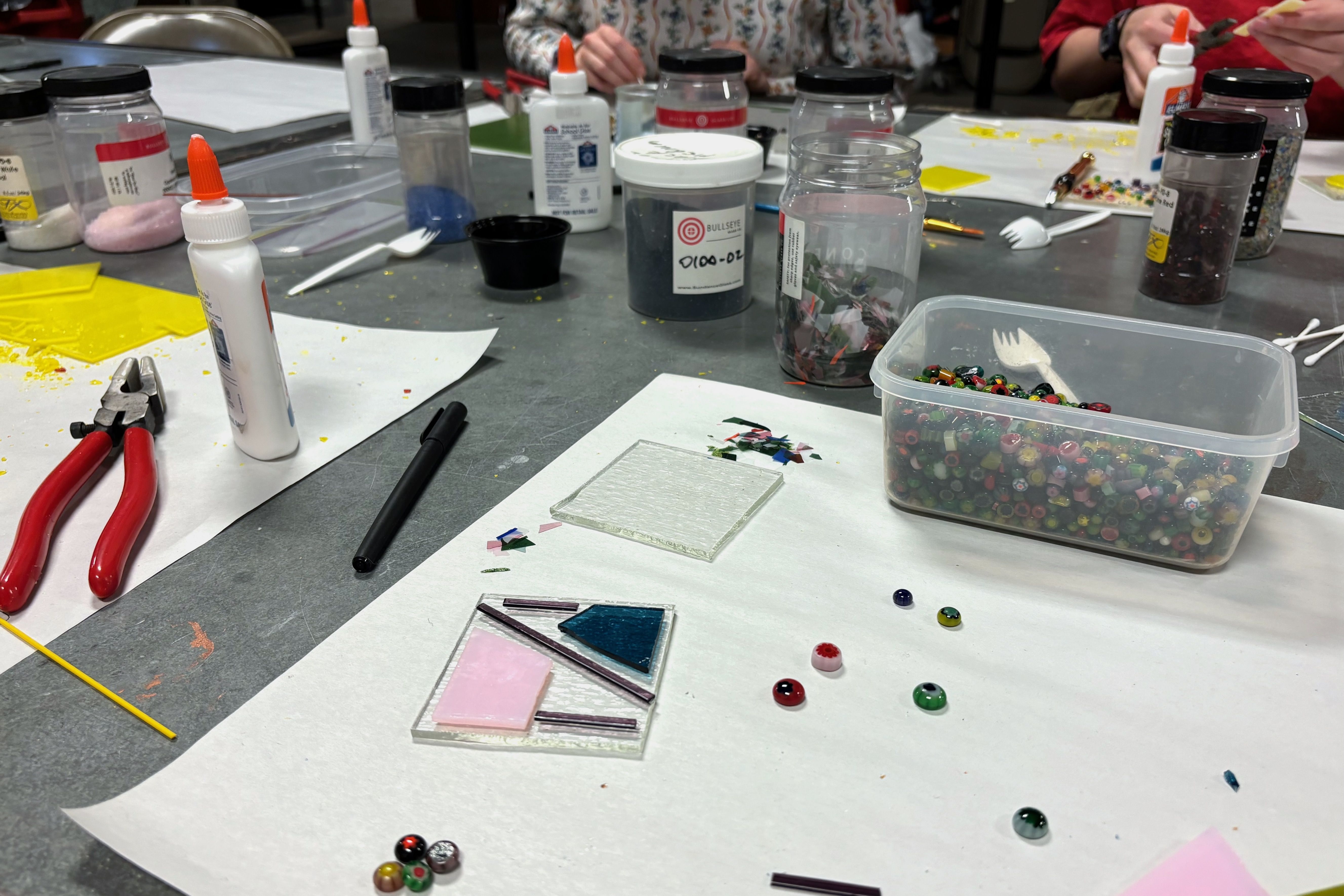 A tabletop full of mosaic-making supplies like beads, glass shards, glue and pliers