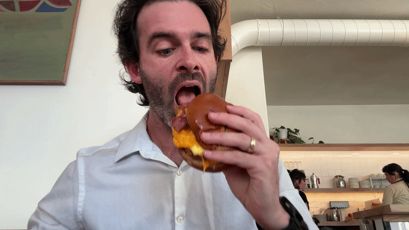 Gif showing reporter Travis in a white shirt biting into a breakfast sandwich as restaurant workers move behind the bar in the background.