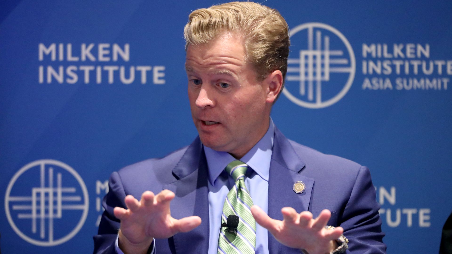 A man with blond hair, gesturing while he speaks, in a light blue business suit, with Milken Institute written on the wall behind him.