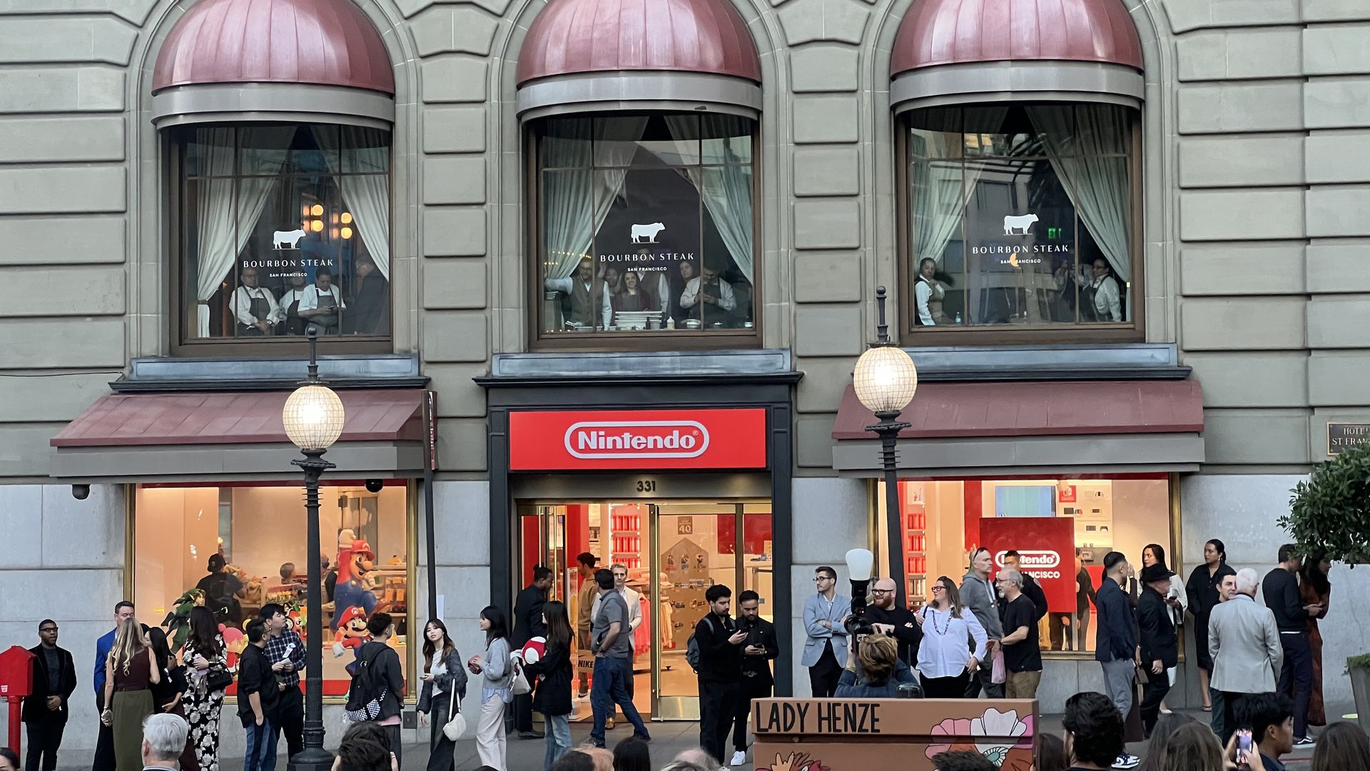 Crowd gathers outside a Nintendo store with a red sign. Above are three arched windows with pink awnings, showing staff inside a restaurant named "Bourbon Steak."