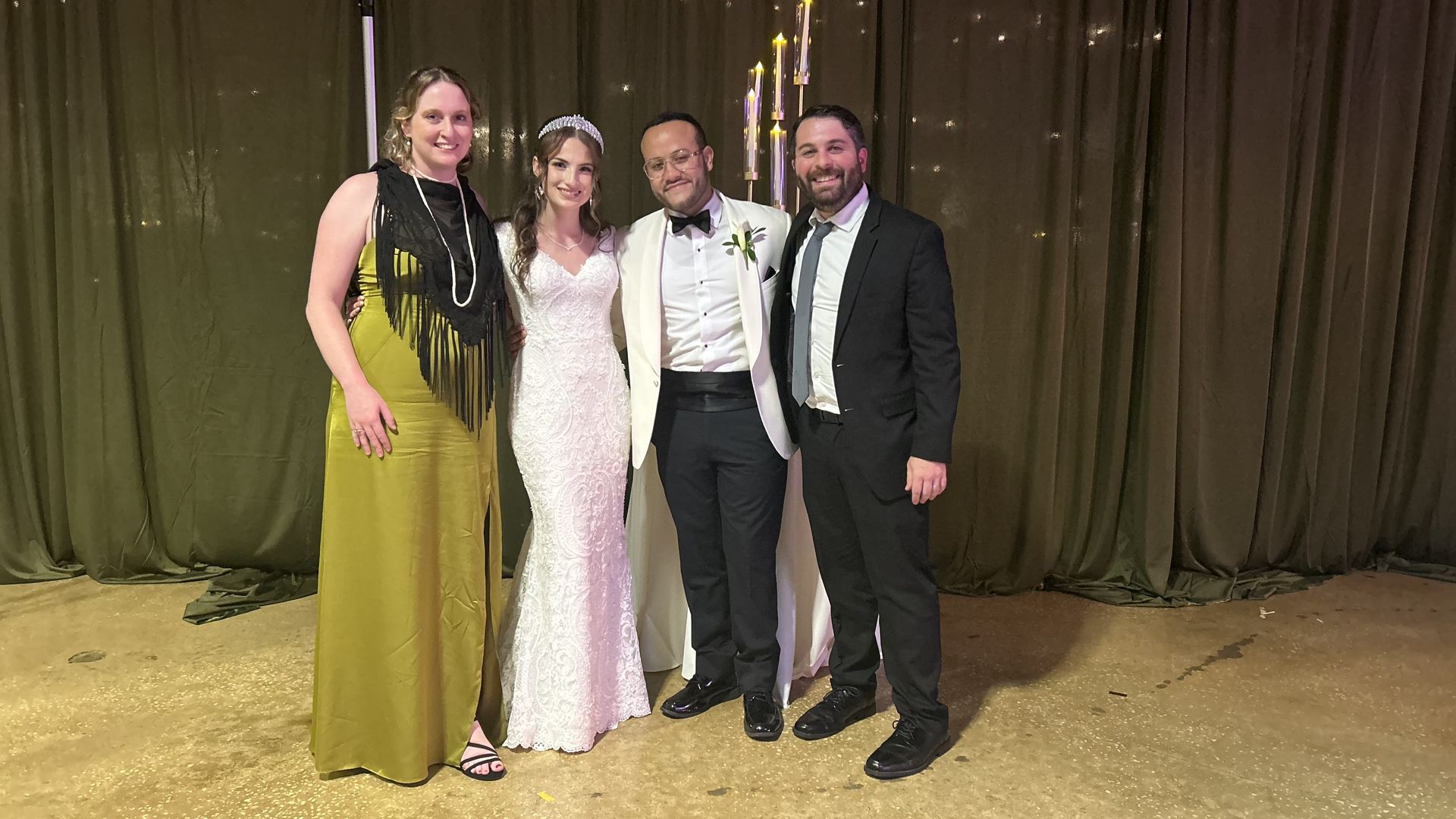 Four people posing indoors at an event with dark curtains behind them. Woman in white lace wedding dress and tiara, man in white tuxedo with bow tie, woman in green dress with black scarf, man in black suit and tie.