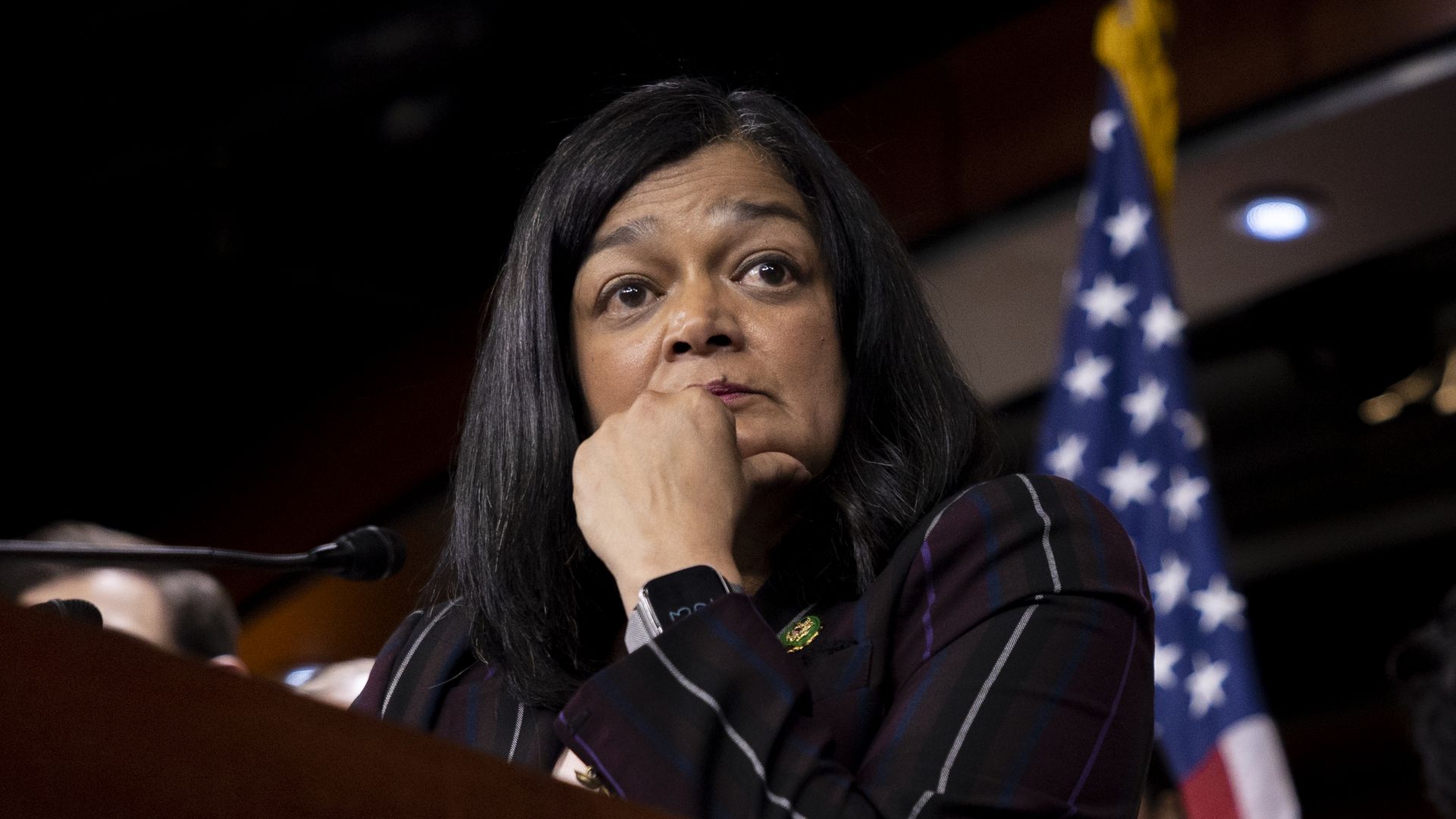 Rep. Pramila Jayapal stands at a podium looking off camera to her left, while resting her elbow on the podium and putting her hand around her chin. 