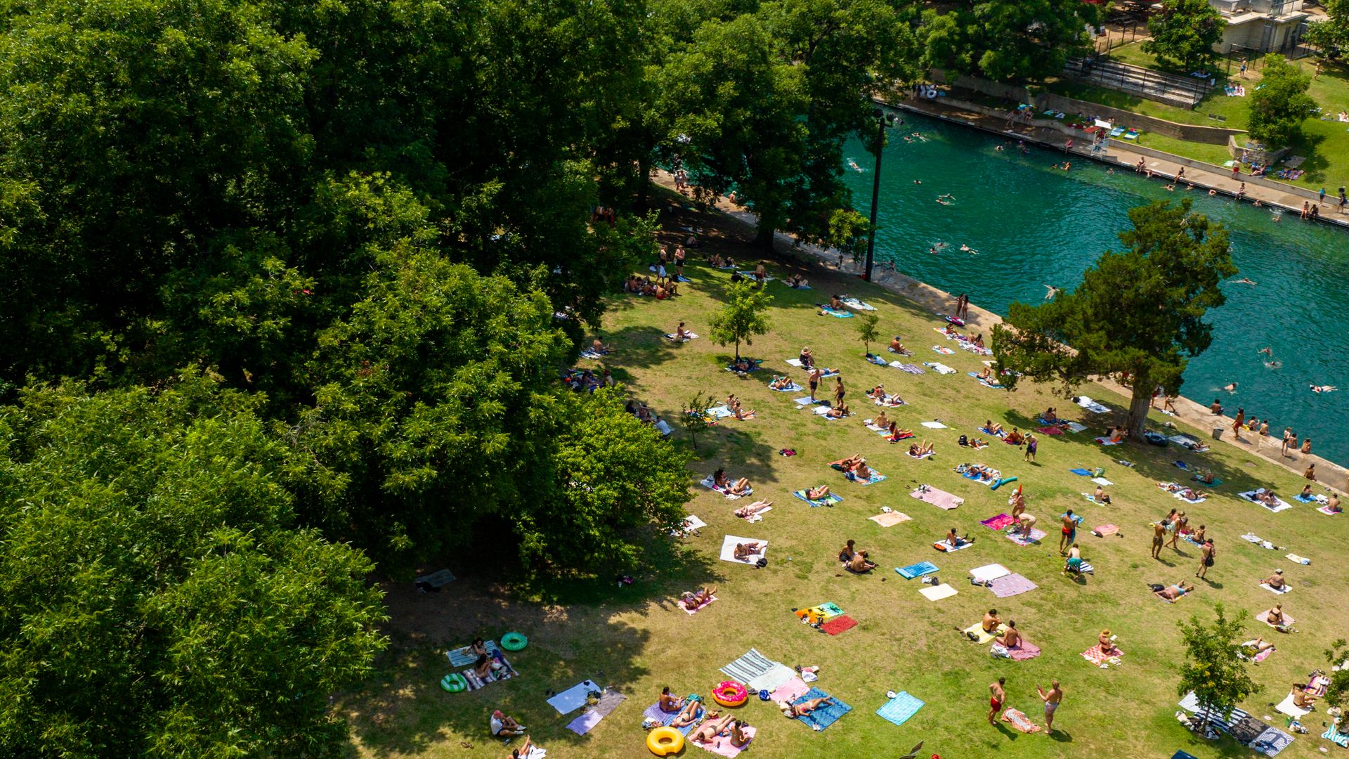 Aerial view of people sunbathing on colorful towels beside a turquoise river lined with trees on a sunny day in a park.