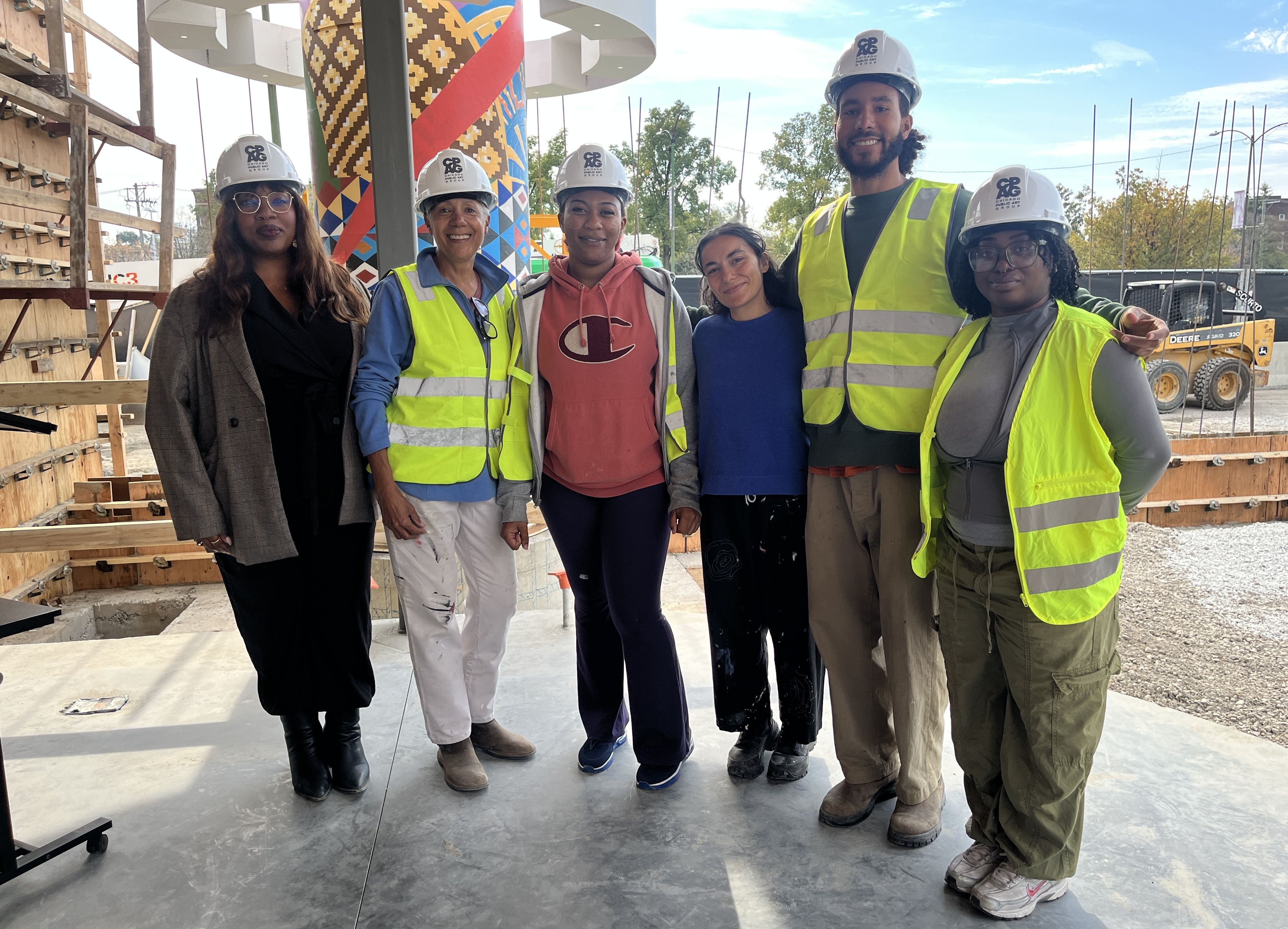 Six people at a construction site, wearing hard hats and high-visibility vests, posing and smiling for the camera. A colorful patterned column and scaffolding stand behind them.