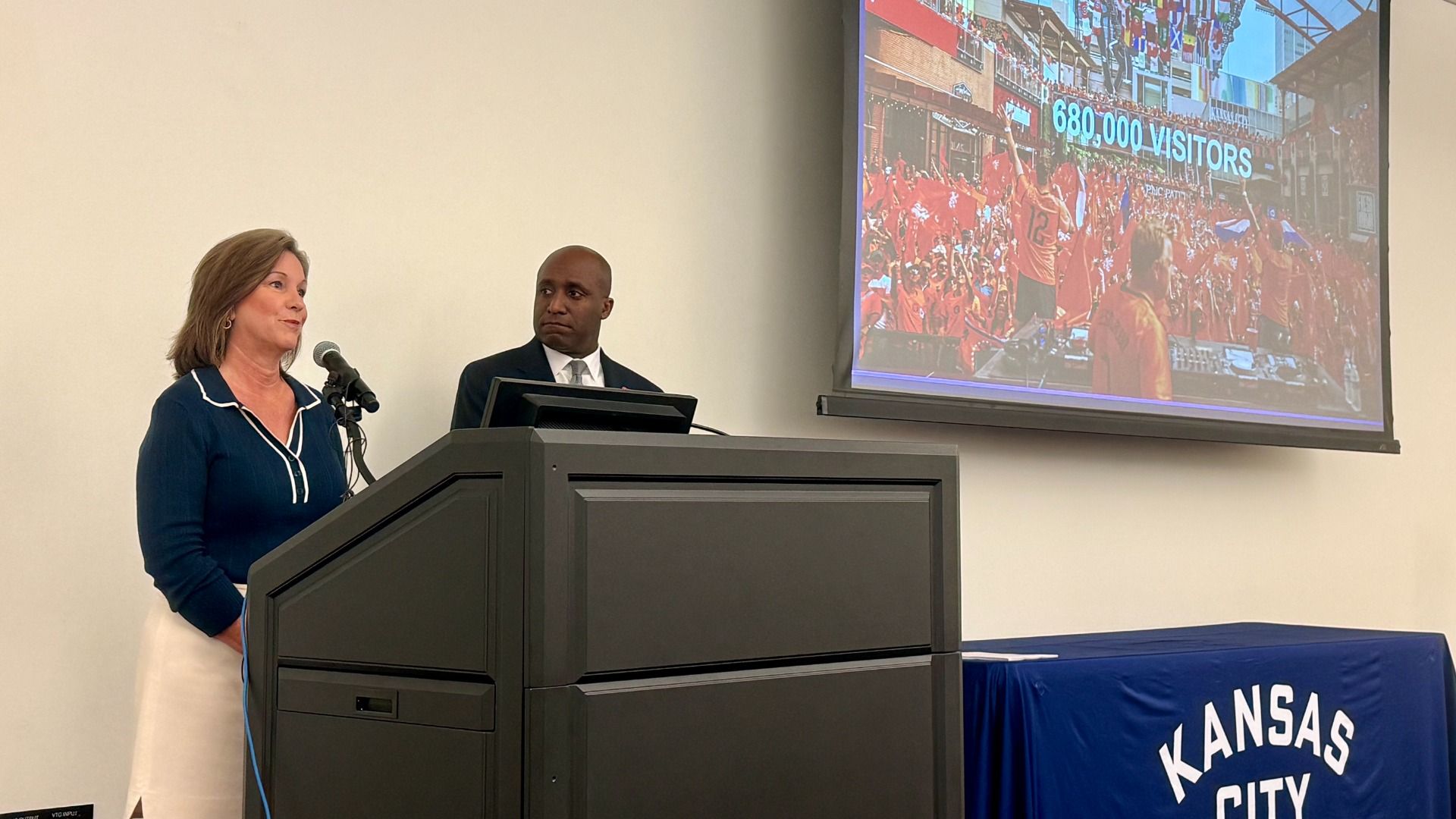 Two speakers at a podium in a conference room: a woman in a blue top and a man in a dark suit, with a large projection screen showing a red-clad crowd and the text 680,000 VISITORS.