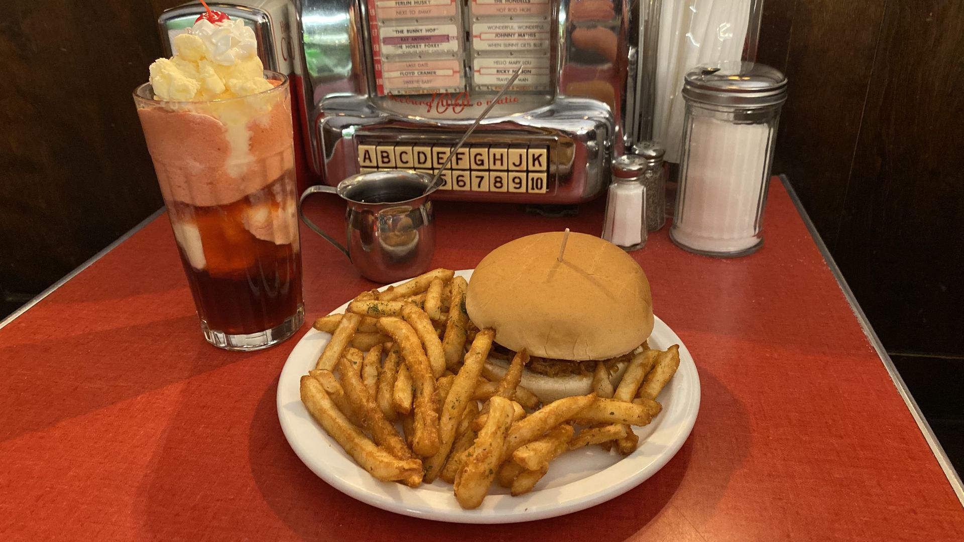 An ice cream soda and a plate with a burger and fries in front of a tabletop jukebox.